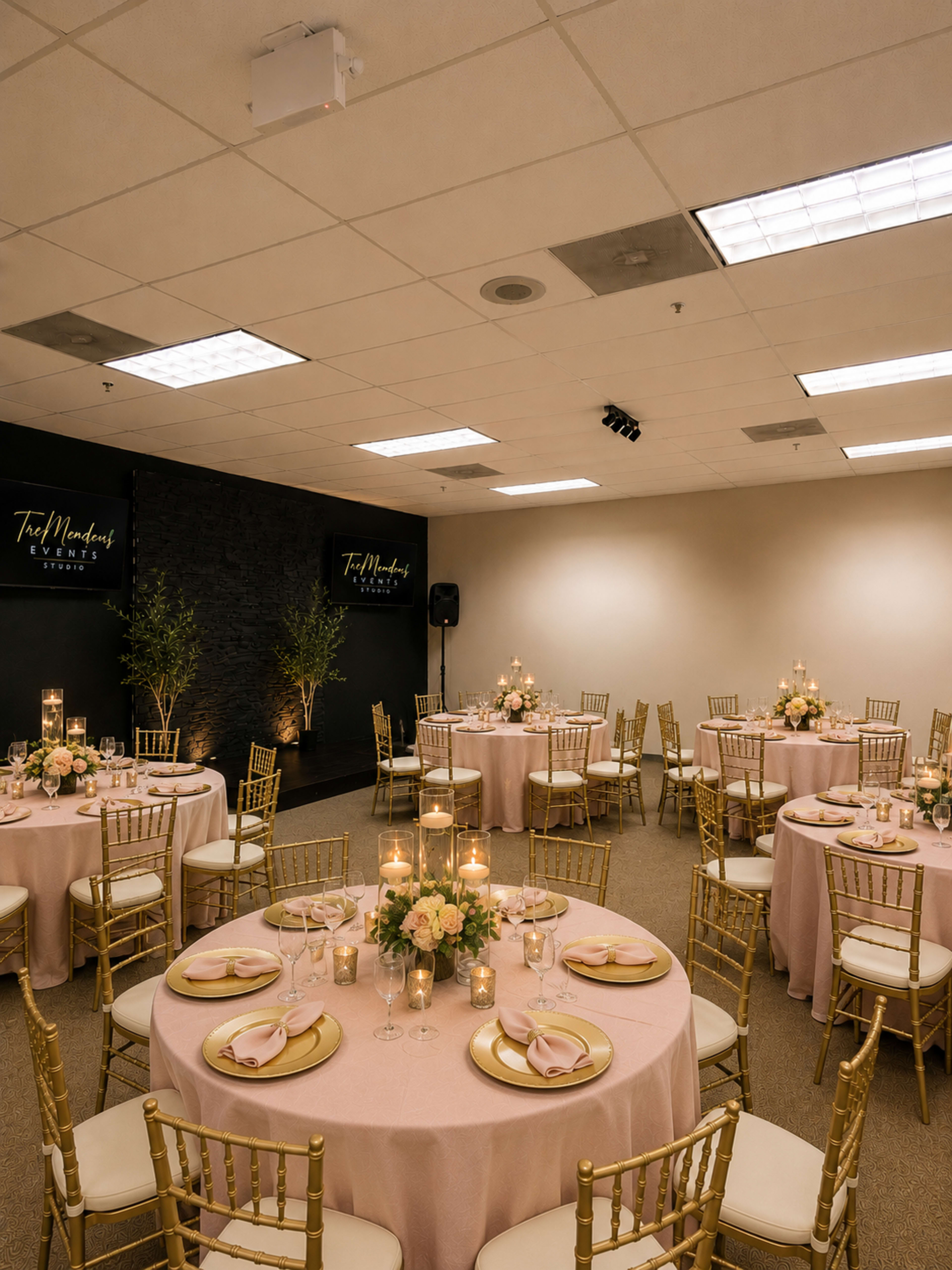 A banquet room is set up with round tables covered in pink tablecloths, gold chairs, and centerpieces of flowers and candles.