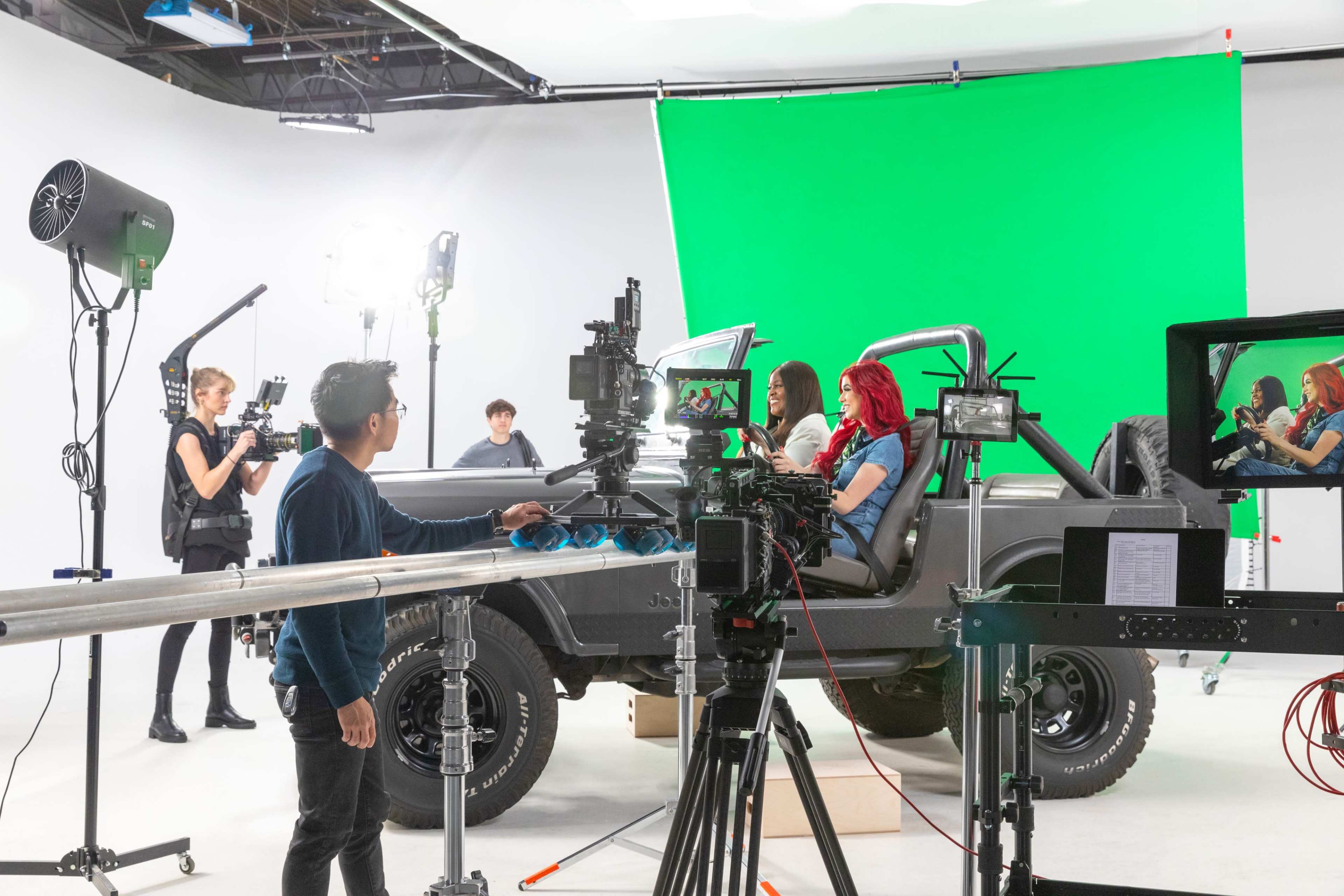 A film crew operates cameras and lighting equipment around a vehicle on a soundstage with a green screen backdrop.