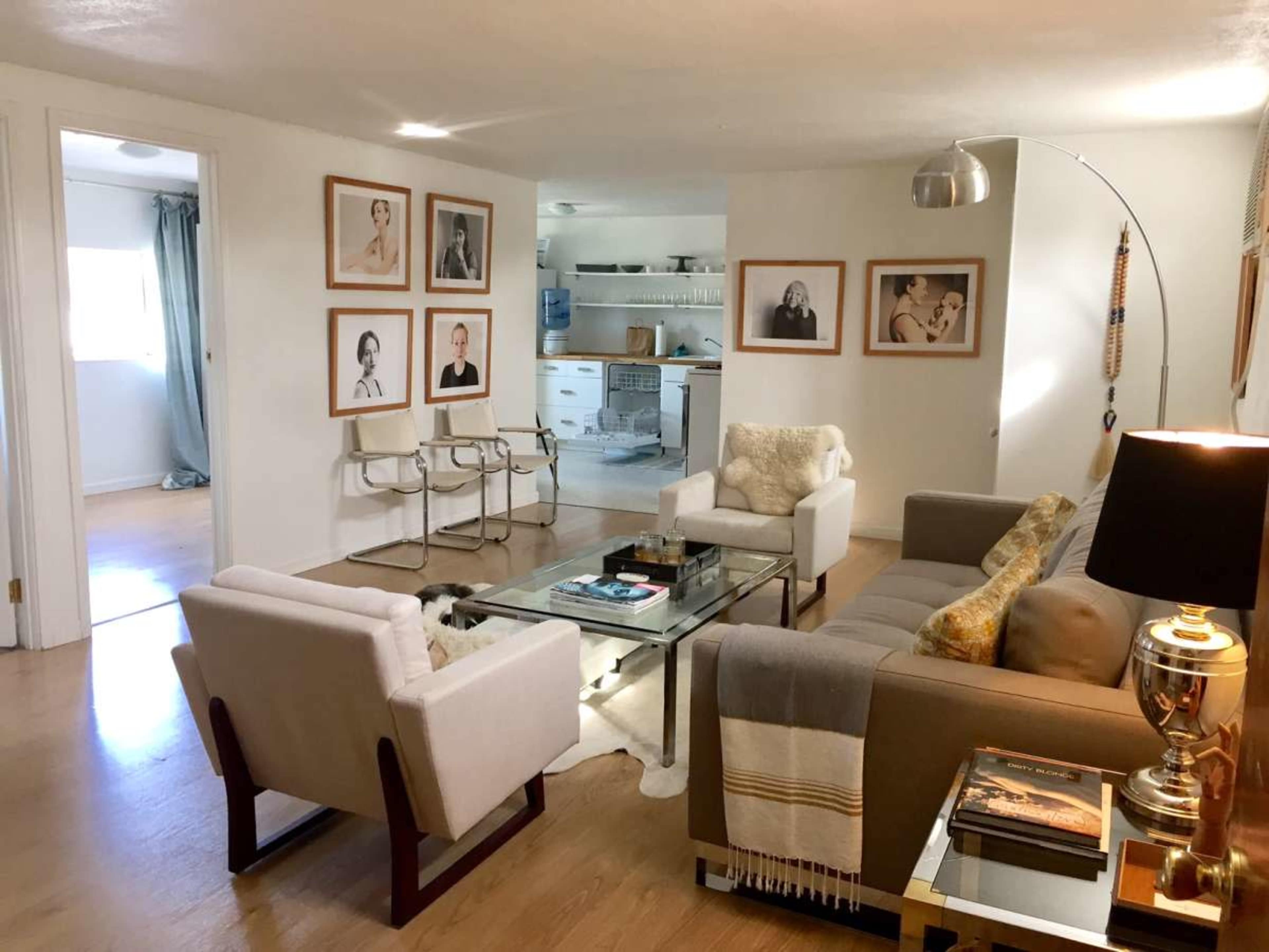 The image shows a modern living room with two white armchairs and a beige sofa, framed black-and-white photographs on the walls, and a glass coffee table in the center.