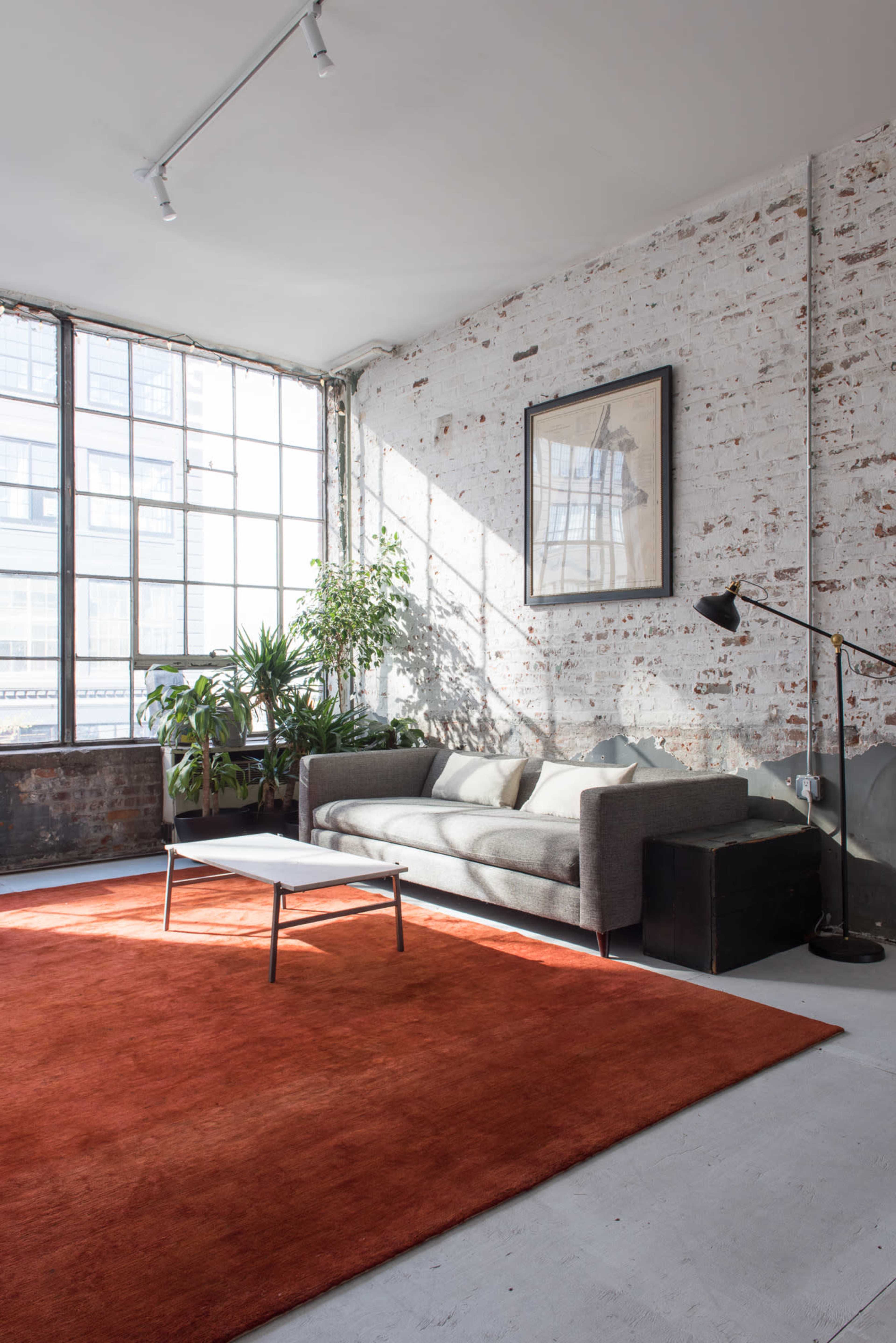 A bright living area features a gray sofa, a coffee table, and a large orange rug, accented by plants and a wall art piece against a brick backdrop.
