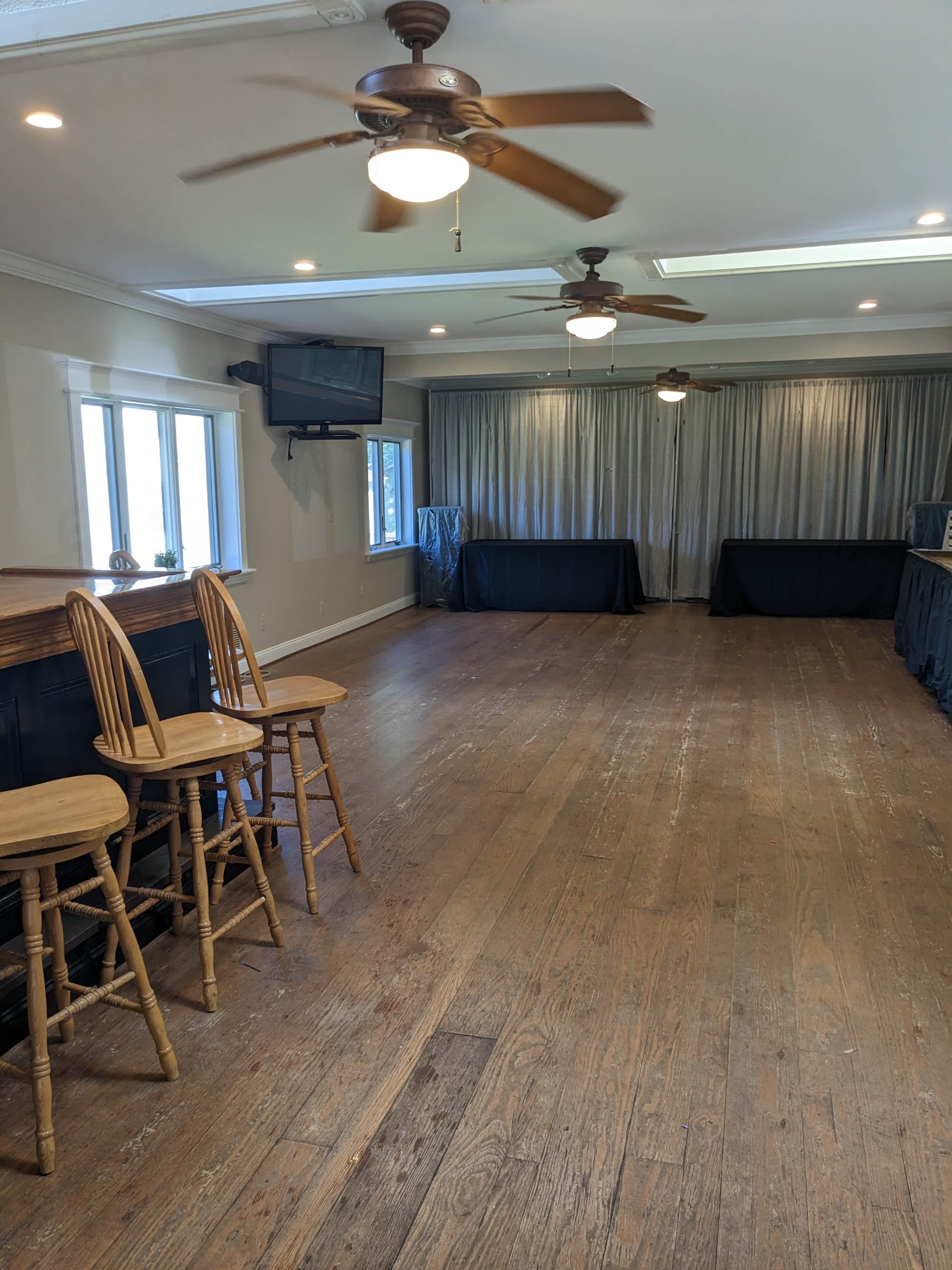 A spacious, empty room with wooden flooring, two ceiling fans, and a bar with three wooden stools on one side.