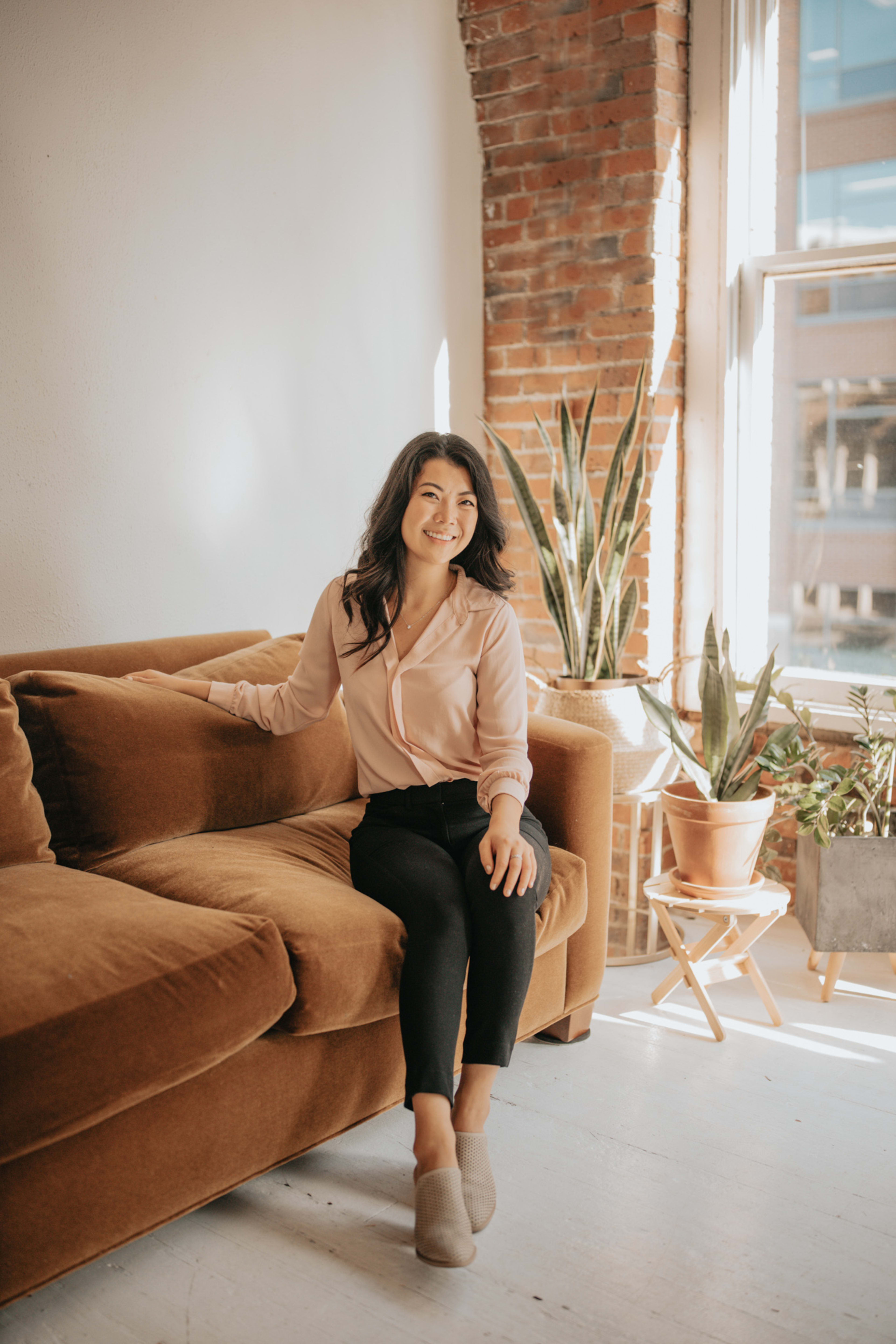 A fashion photo shoot with a woman sitting on top of a brown couch surrounded by plants.