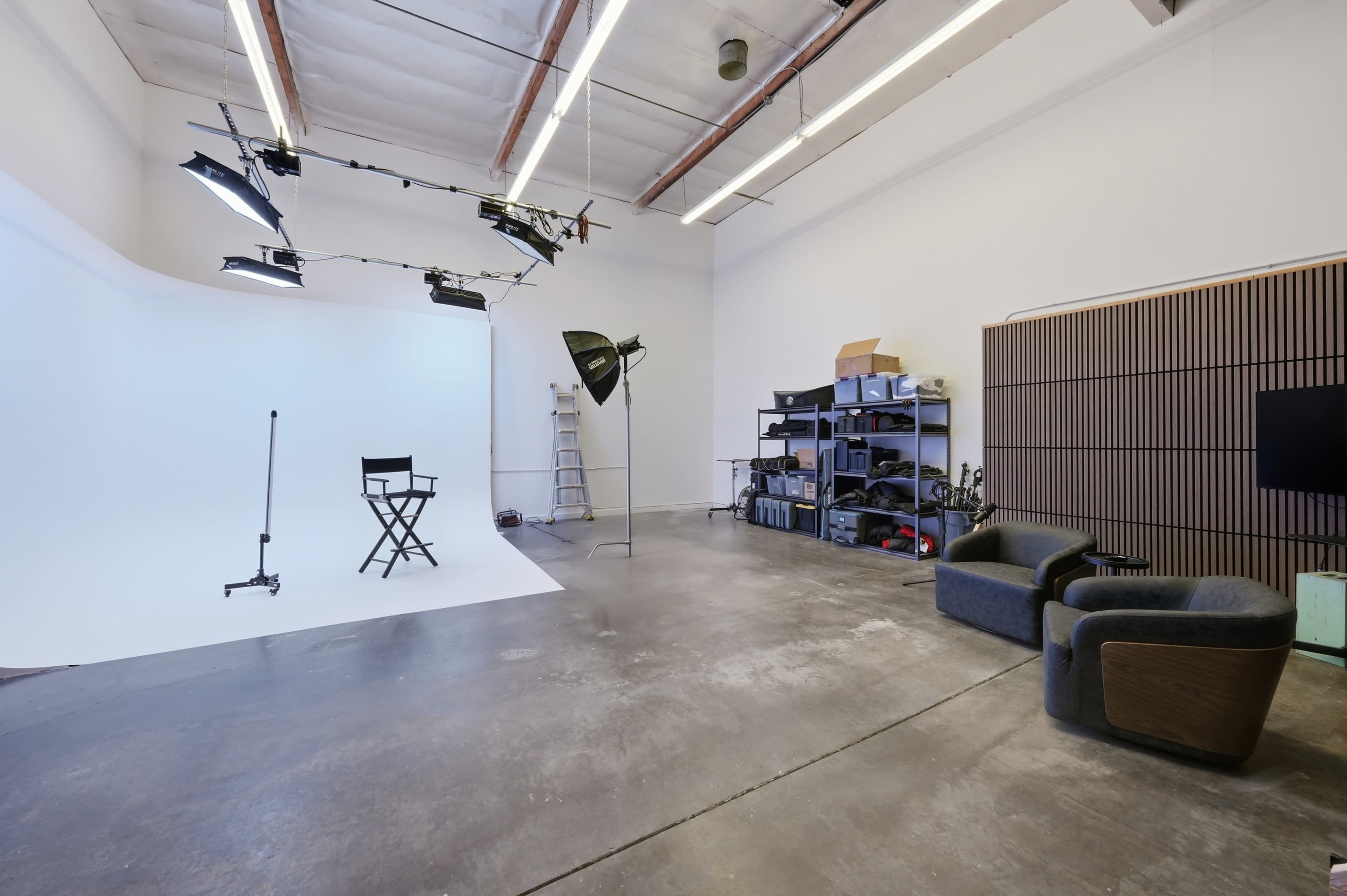 The image shows a photography studio featuring a white backdrop, various lighting equipment, a director's chair, and storage shelves filled with equipment.