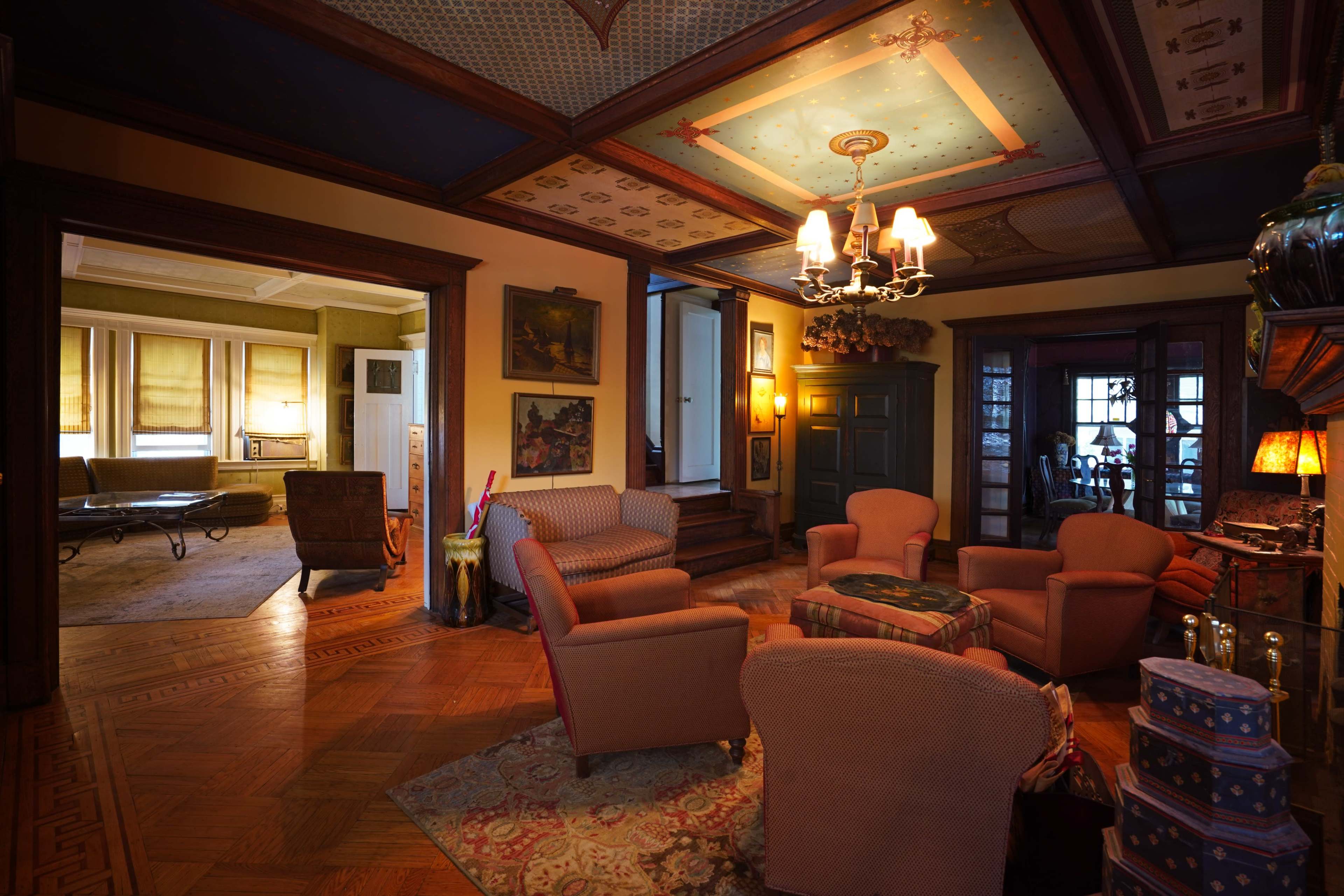 A spacious living room with four upholstered chairs arranged around a small table, featuring decorative ceilings and wooden accents, leading to another room visible in the background.