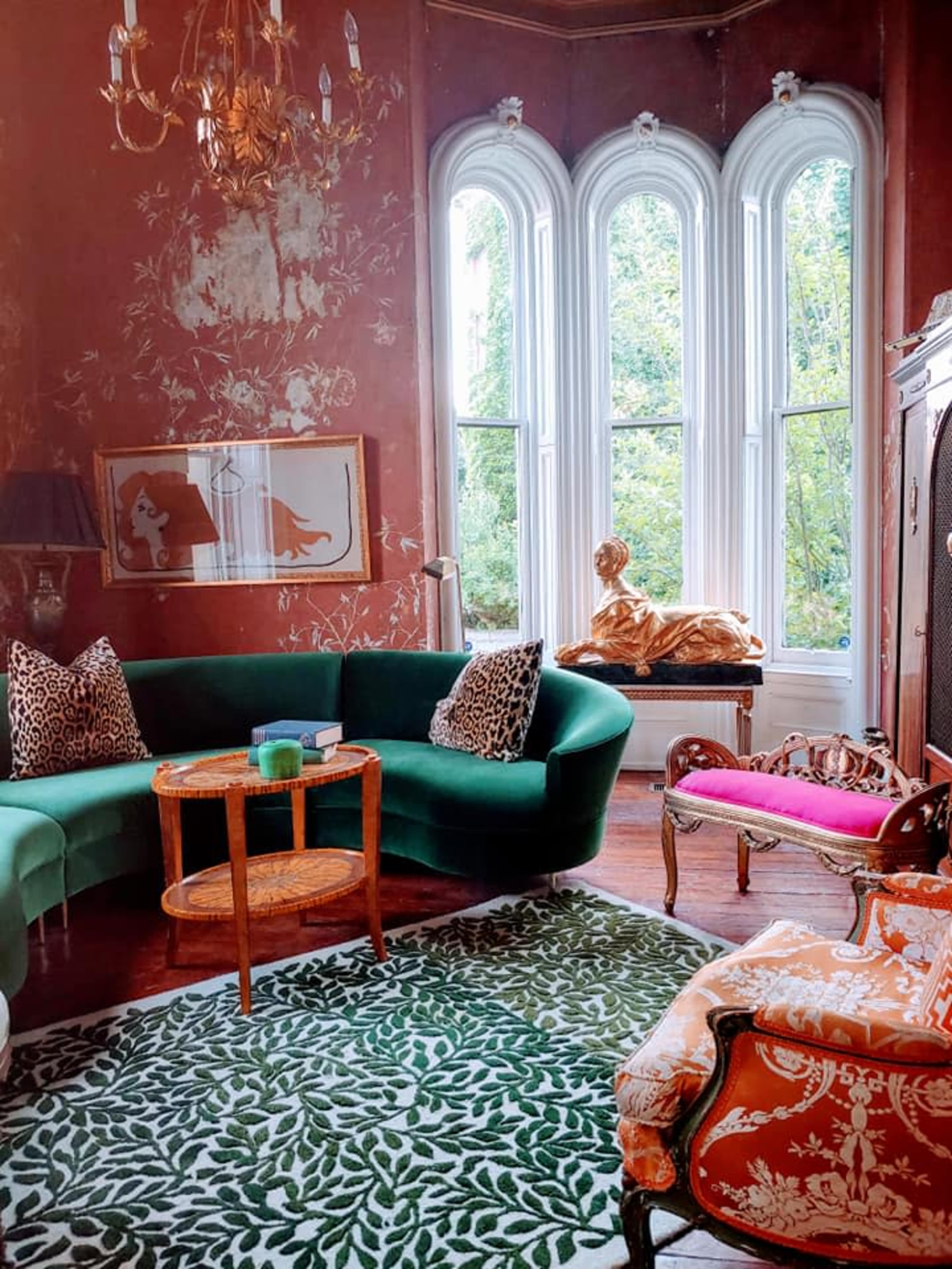 The image shows a decorated sitting room with a green curved sofa, a wooden side table, and ornate chairs, all accented by large windows and a golden statue in the corner.