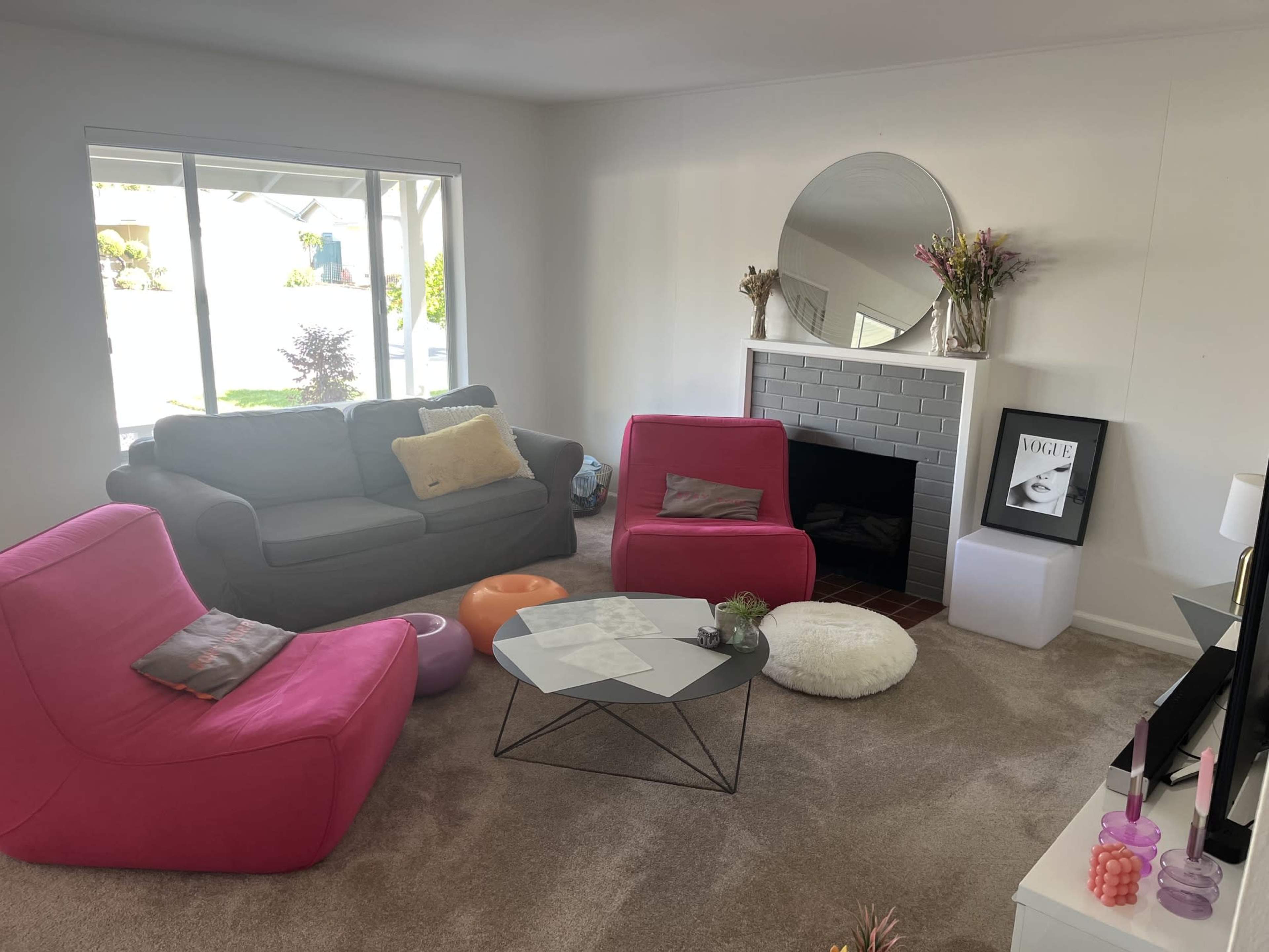 The image shows a cozy living room with a gray sofa, two pink lounge chairs, a glass coffee table, and a fireplace with a round mirror above it.
