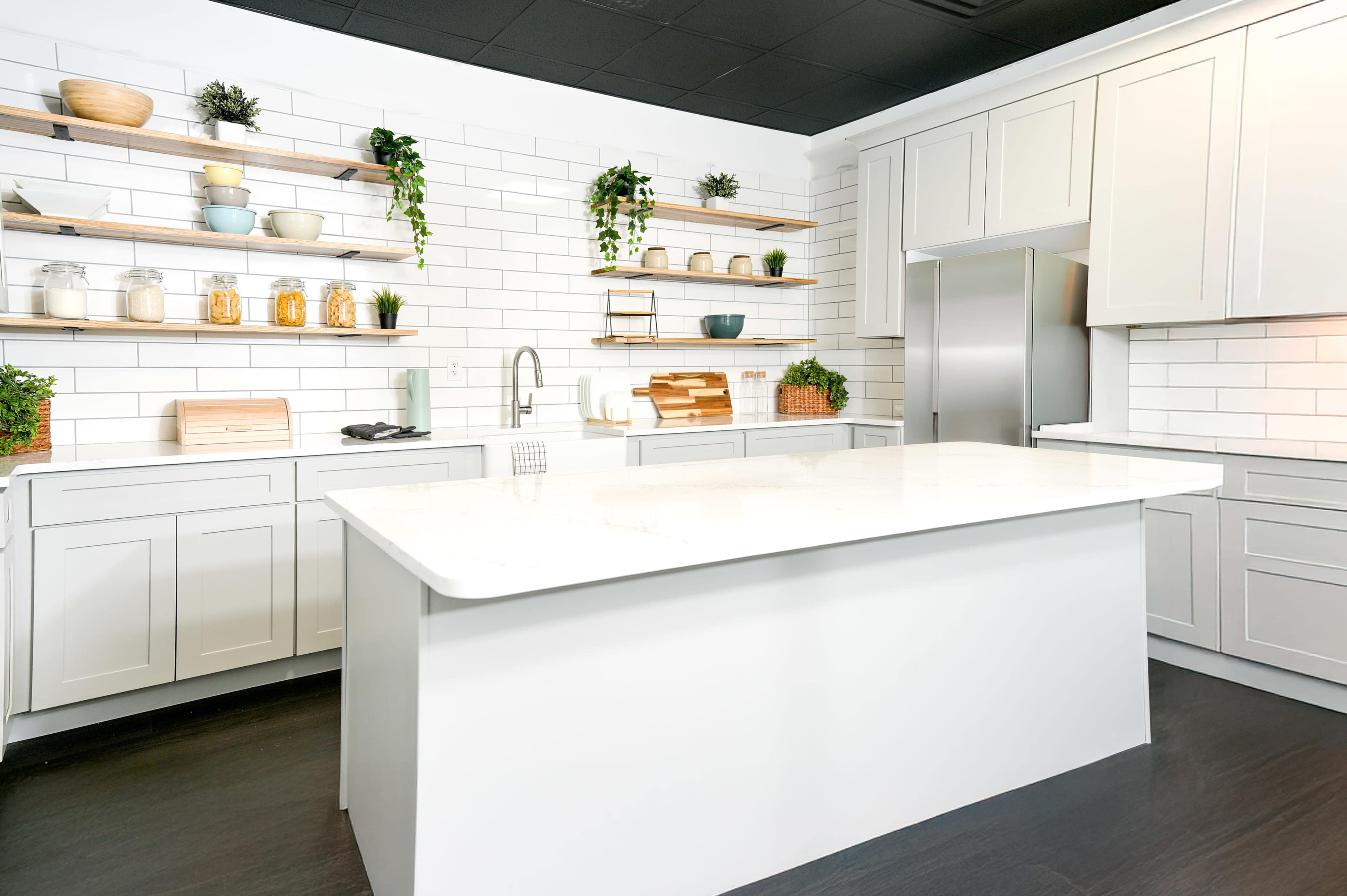 The image shows a modern kitchen featuring a white island, light gray cabinetry, open shelving with decorative items, and a tiled back wall.