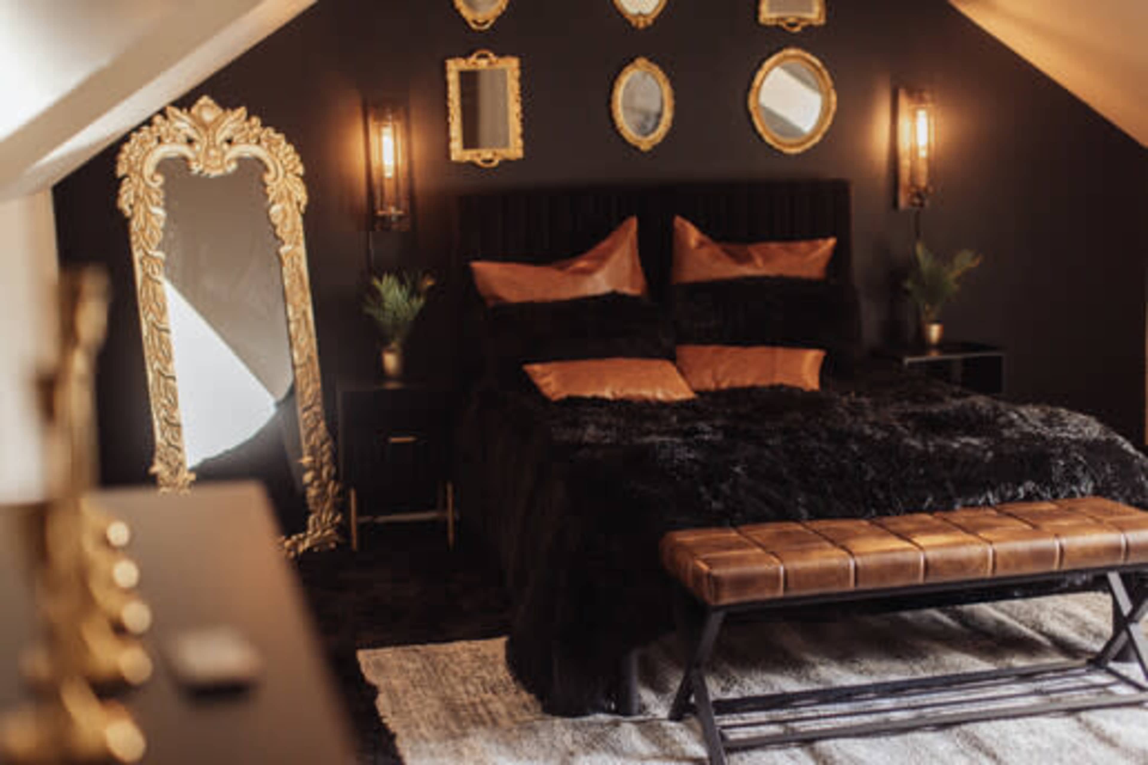 The image shows a stylish bedroom with a large bed covered in a black fur blanket, flanked by decorative mirrors on the walls and a bench at the foot of the bed.