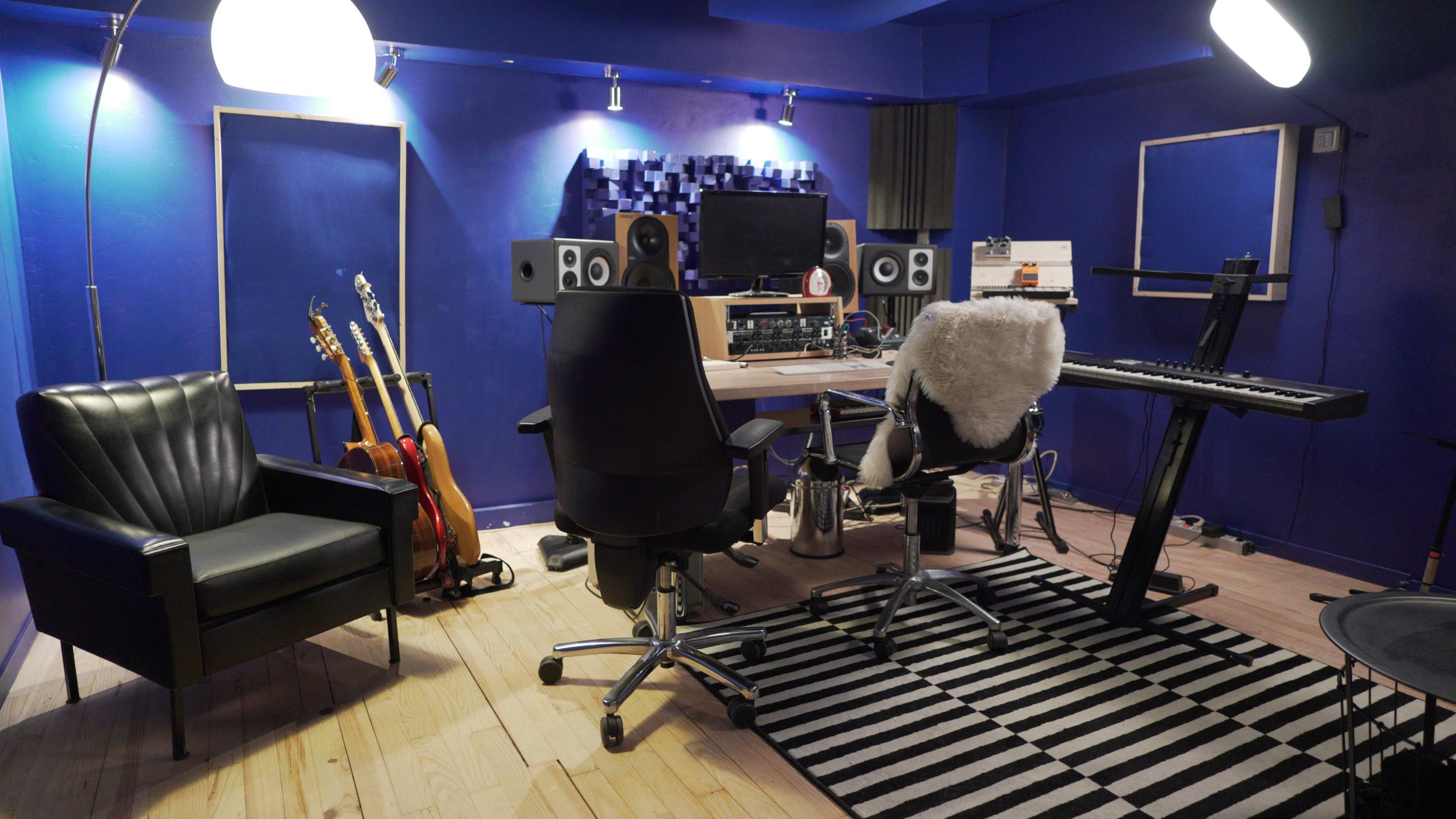 A music studio features a desk with recording equipment, a keyboard, and a chair, surrounded by guitars and illuminated by overhead lights against blue walls.
