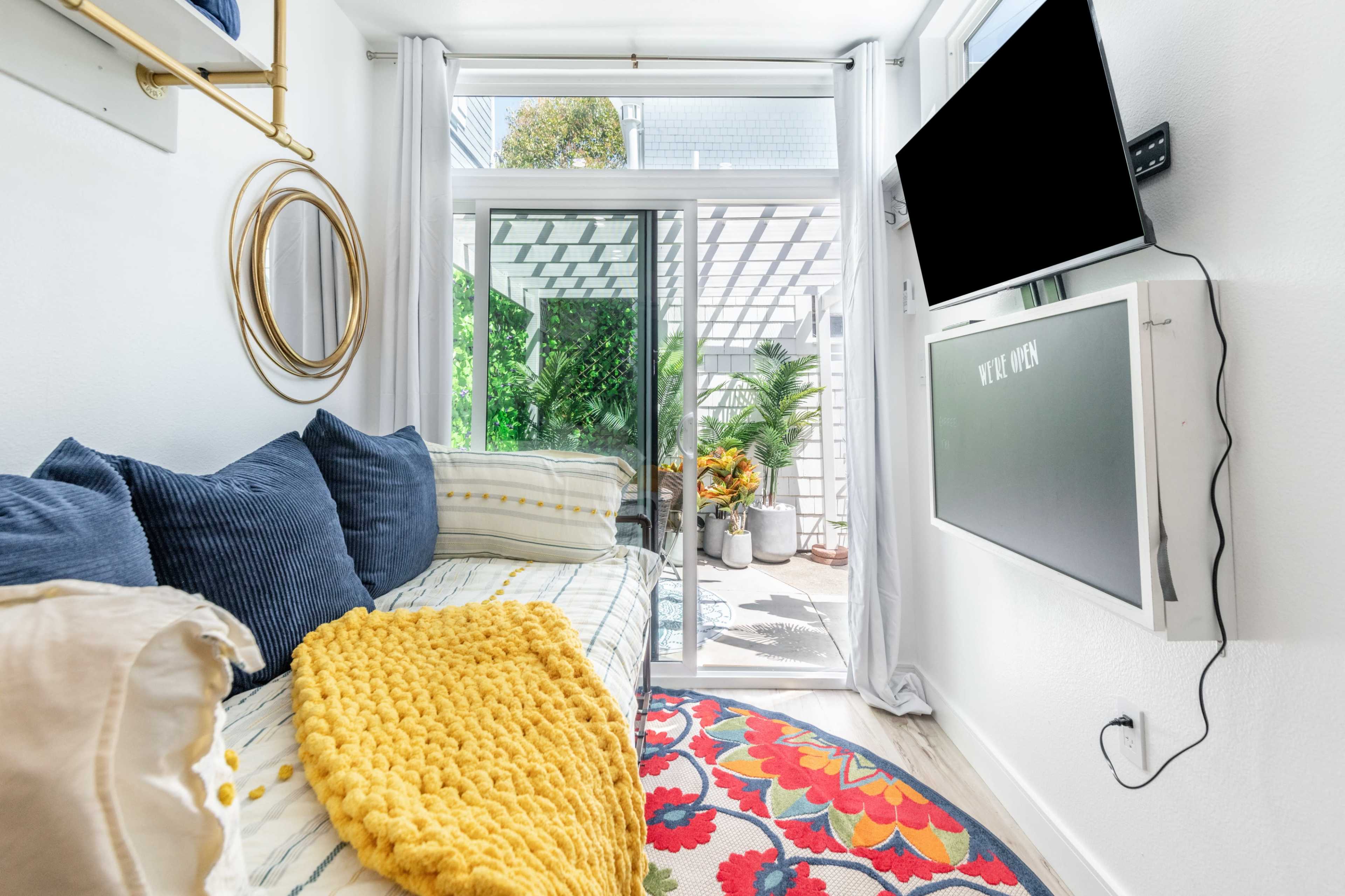 A bright living space features a sofa adorned with a yellow blanket, a colorful rug, and a mounted television, overlooking a sunny outdoor area through glass doors.