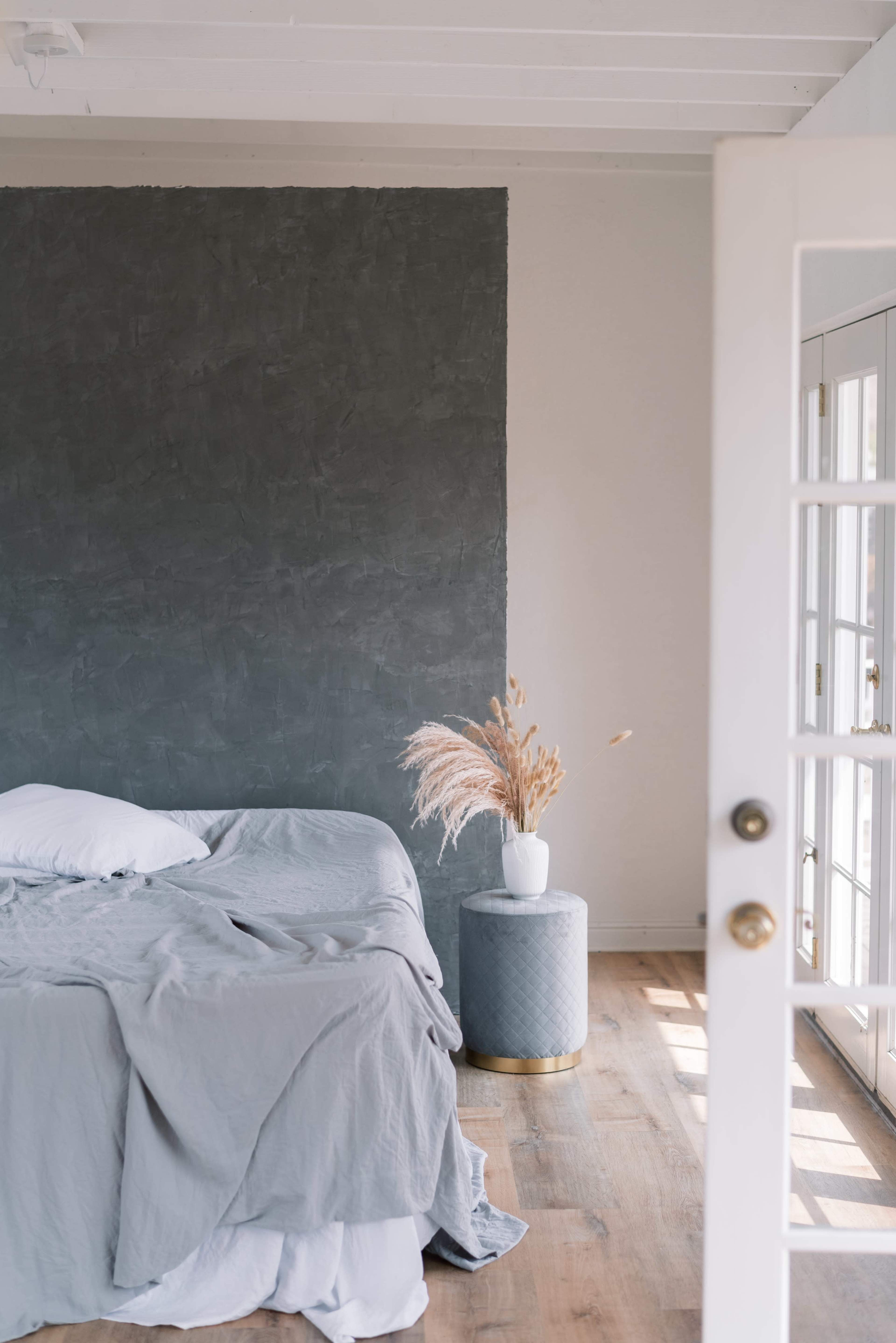 A bedroom features a bed with a gray blanket, a round blue side table with a white vase containing dried plants, and a large window allowing natural light.