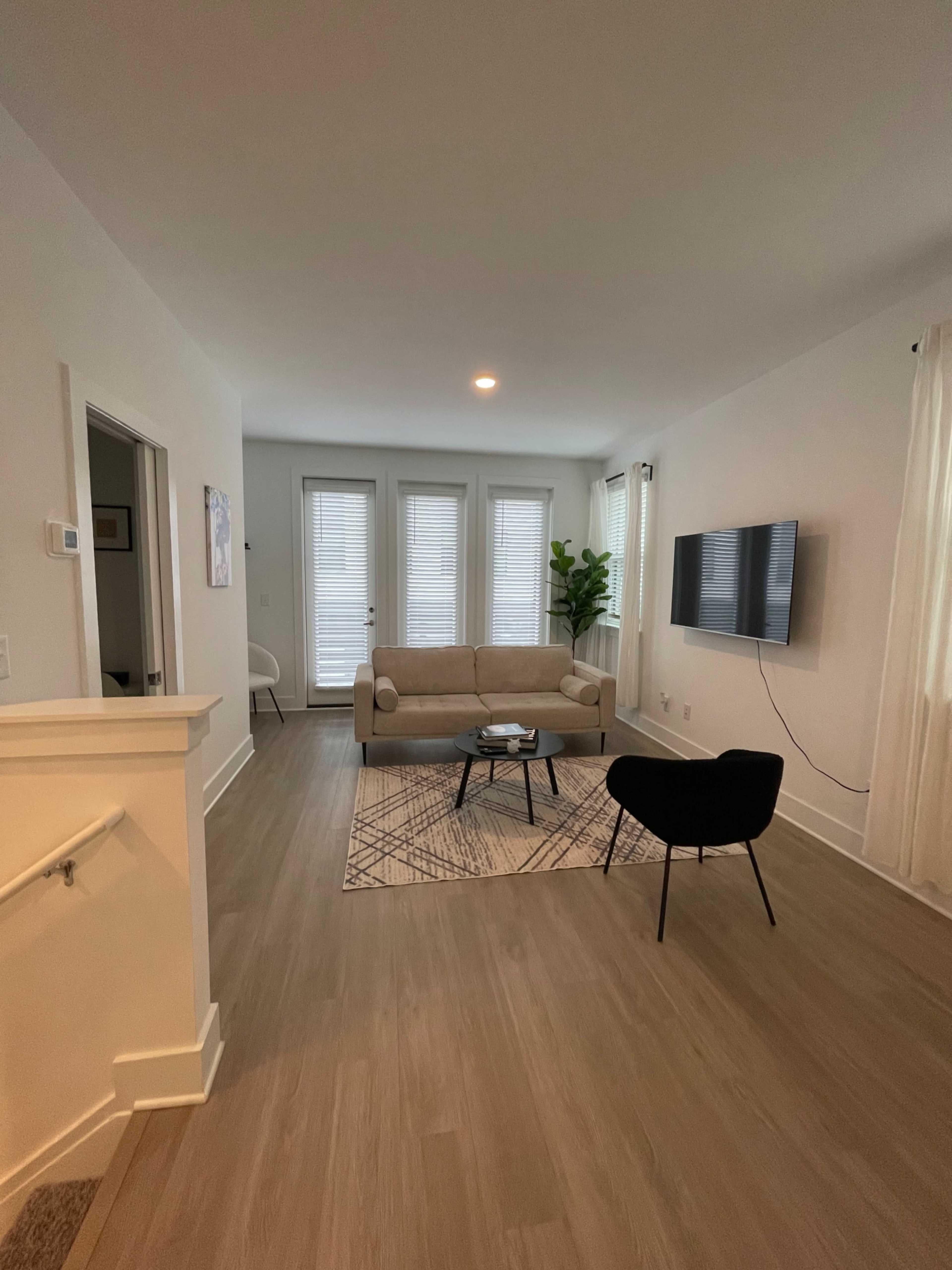 The image shows a modern living room with a light-colored sofa, a black chair, a coffee table on a patterned rug, and large windows with closed blinds.