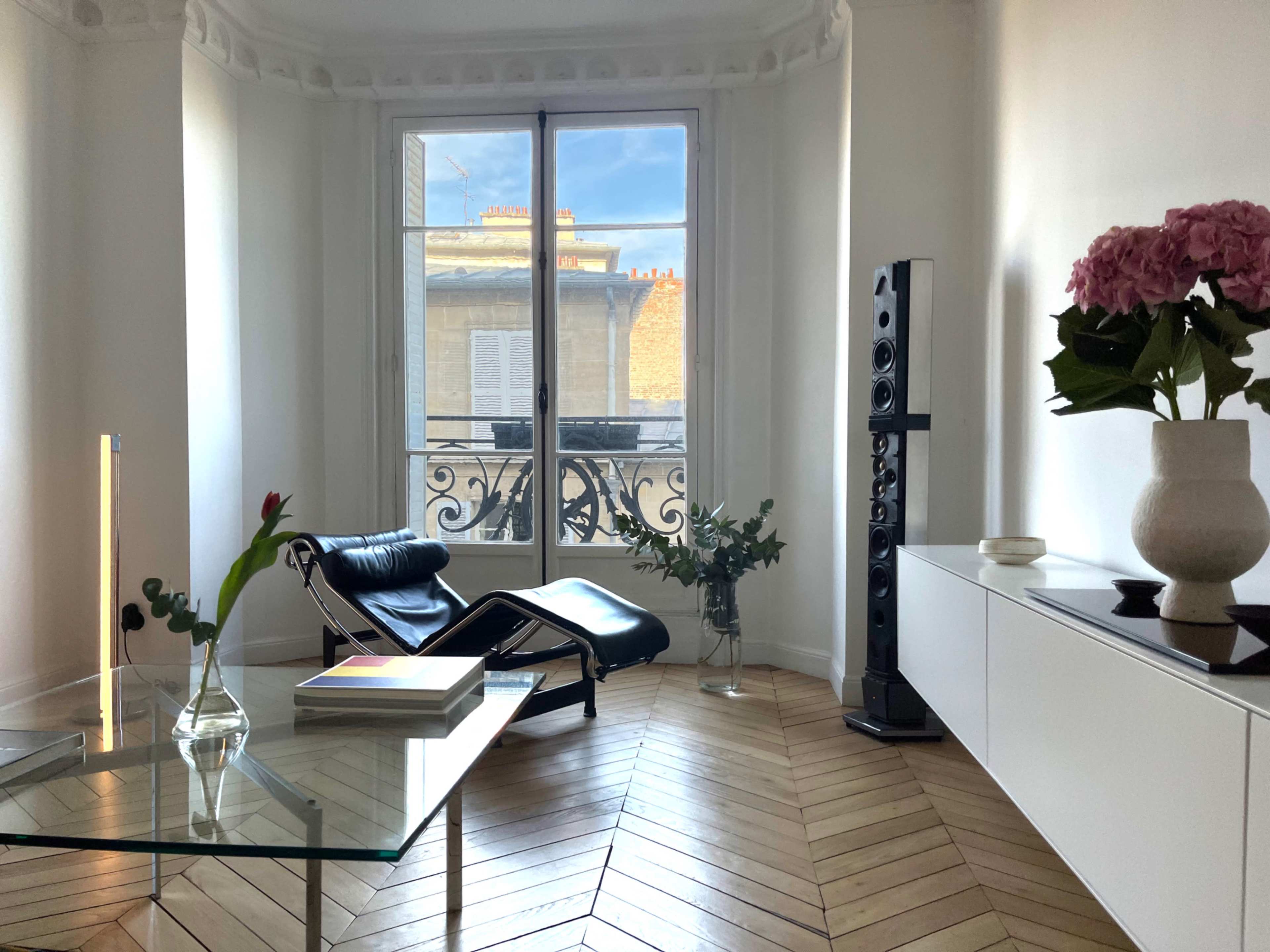 The image shows a minimalistic living room with a large window, elegant wooden flooring, a lounge chair, a glass table, and modern decor elements.