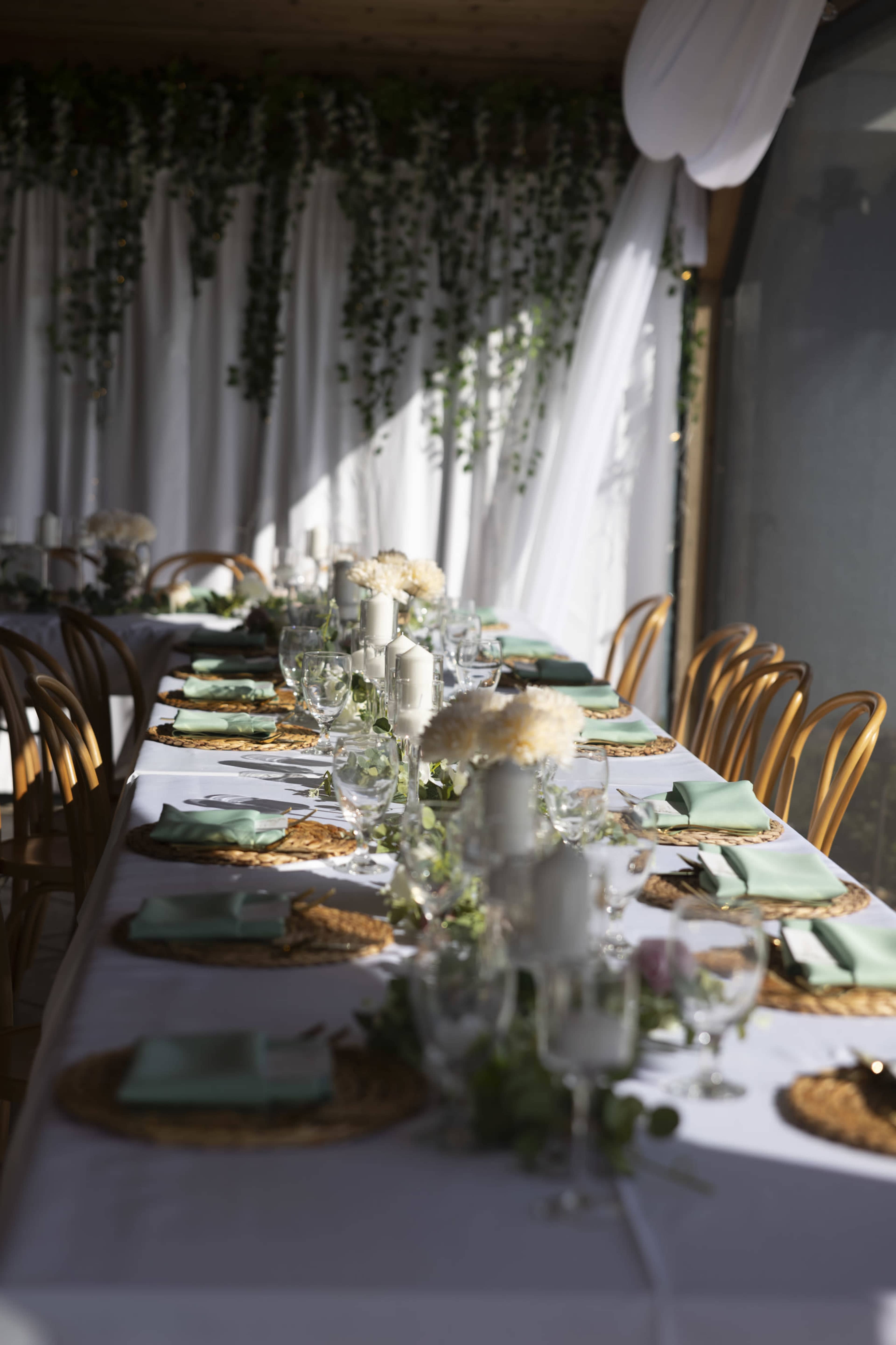 The image shows a long dining table set with various tableware, glasses, and centerpieces, decorated with greenery and flowers against a backdrop of draped fabric.