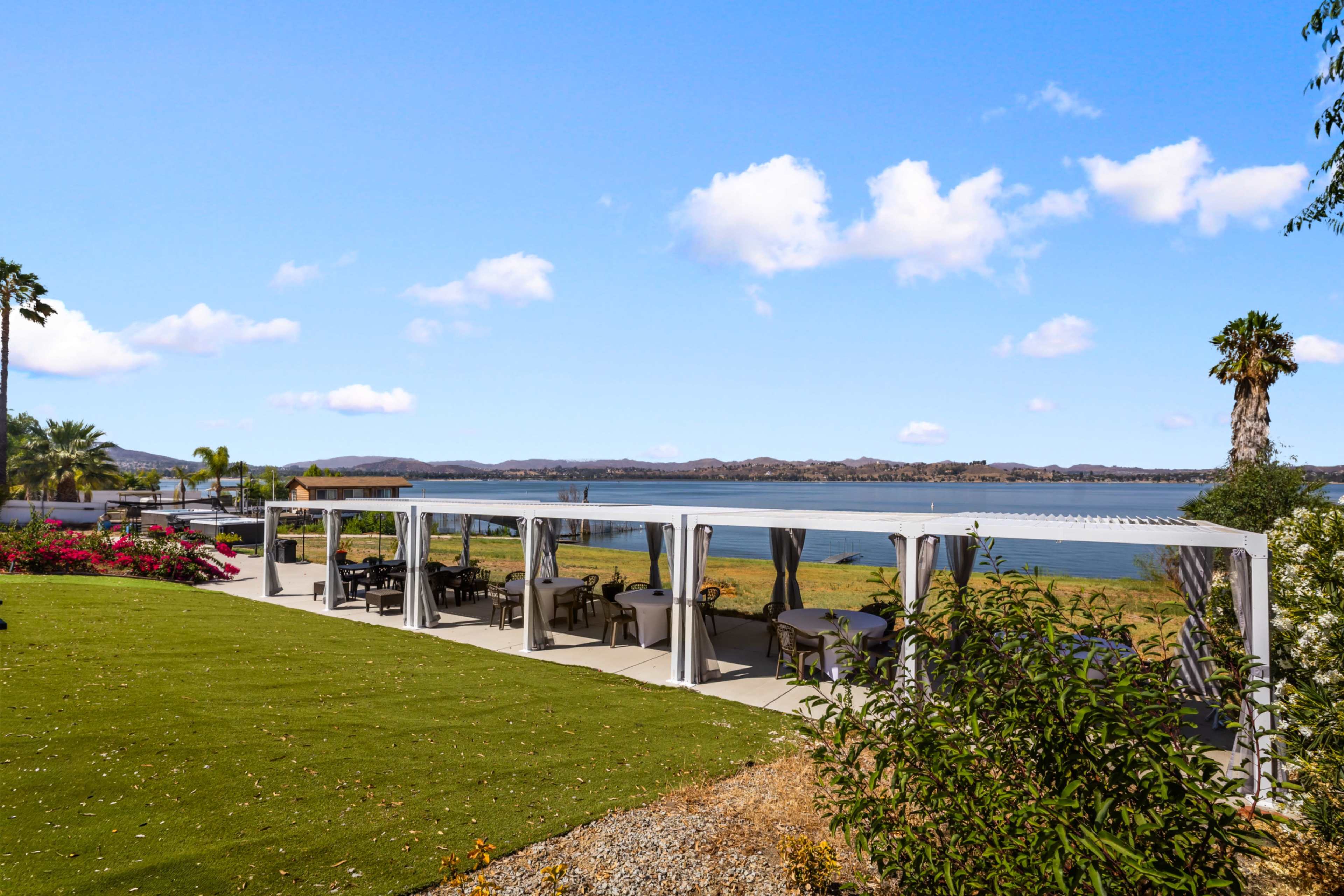 The image shows a lakeside patio with several covered seating areas and a view of the water, surrounded by greenery and shrubs.