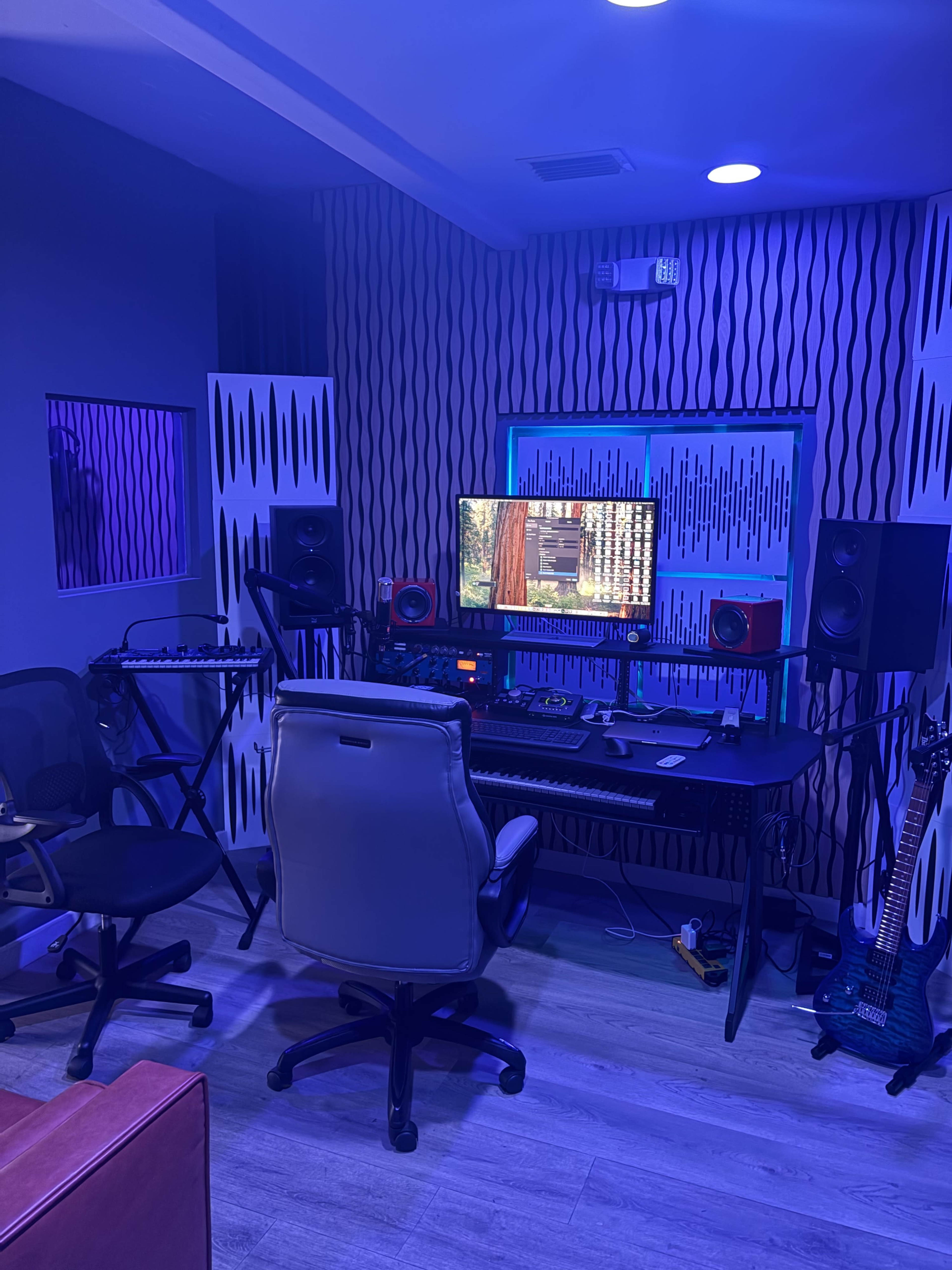 The image shows a modern music studio with a desk featuring a computer screen, audio monitors, and various musical equipment, all illuminated by blue lighting.