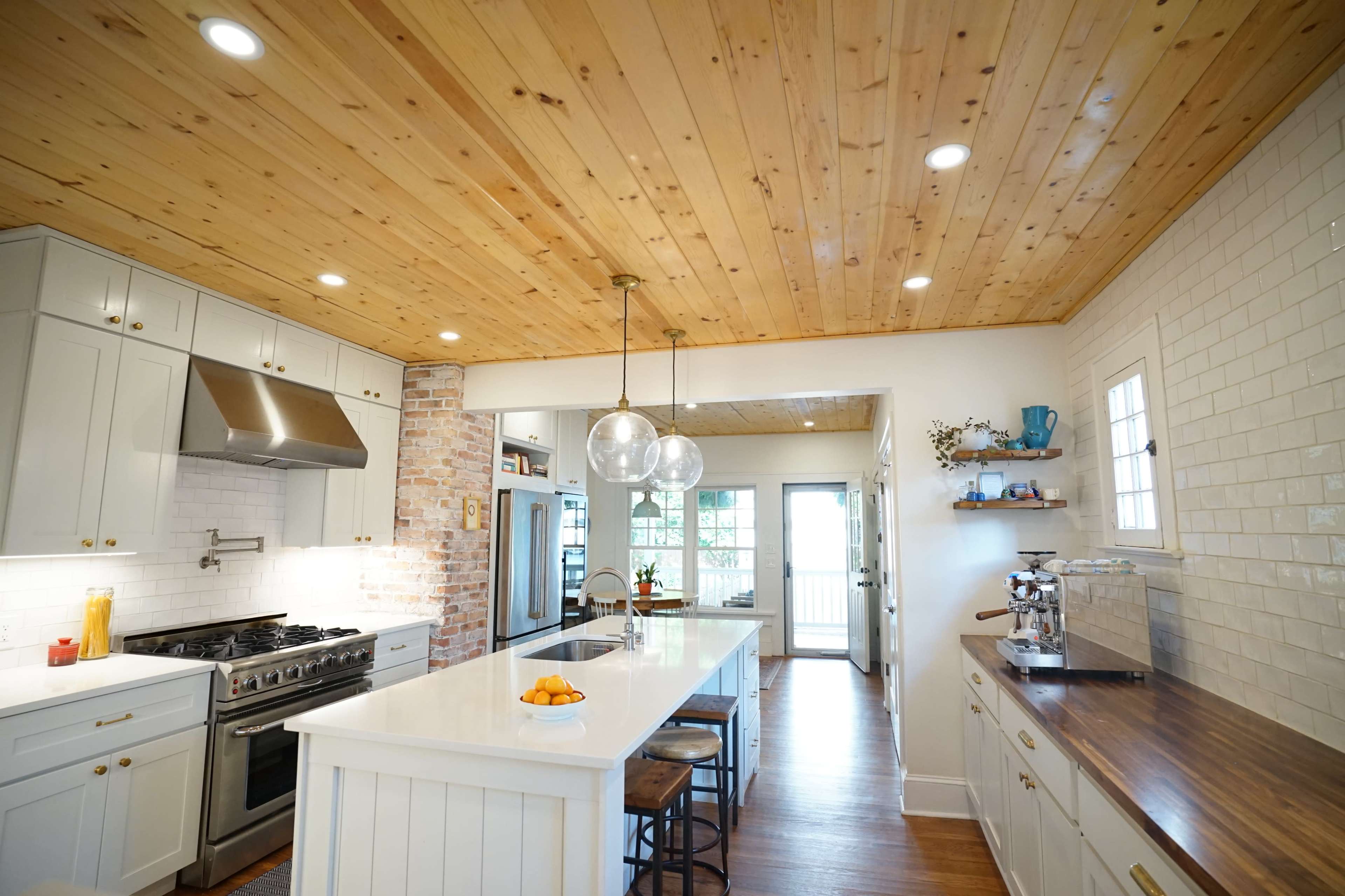 A bright kitchen features a large island with a white countertop, stainless steel appliances, and wooden accents including a ceiling and countertop.