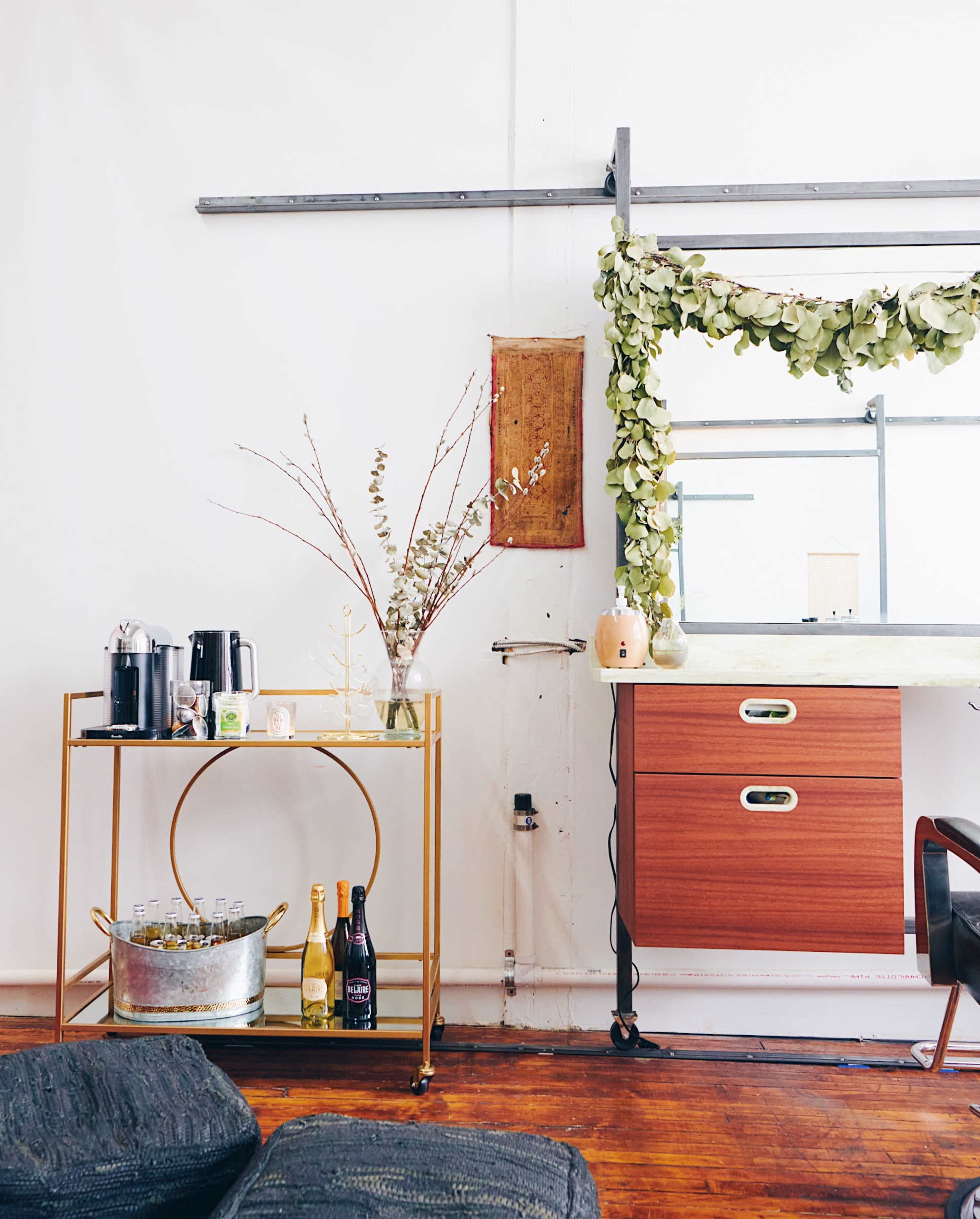 A gold metal bar cart holds coffee, a flower vase, and bottles, next to a wooden cabinet adorned with greenery.