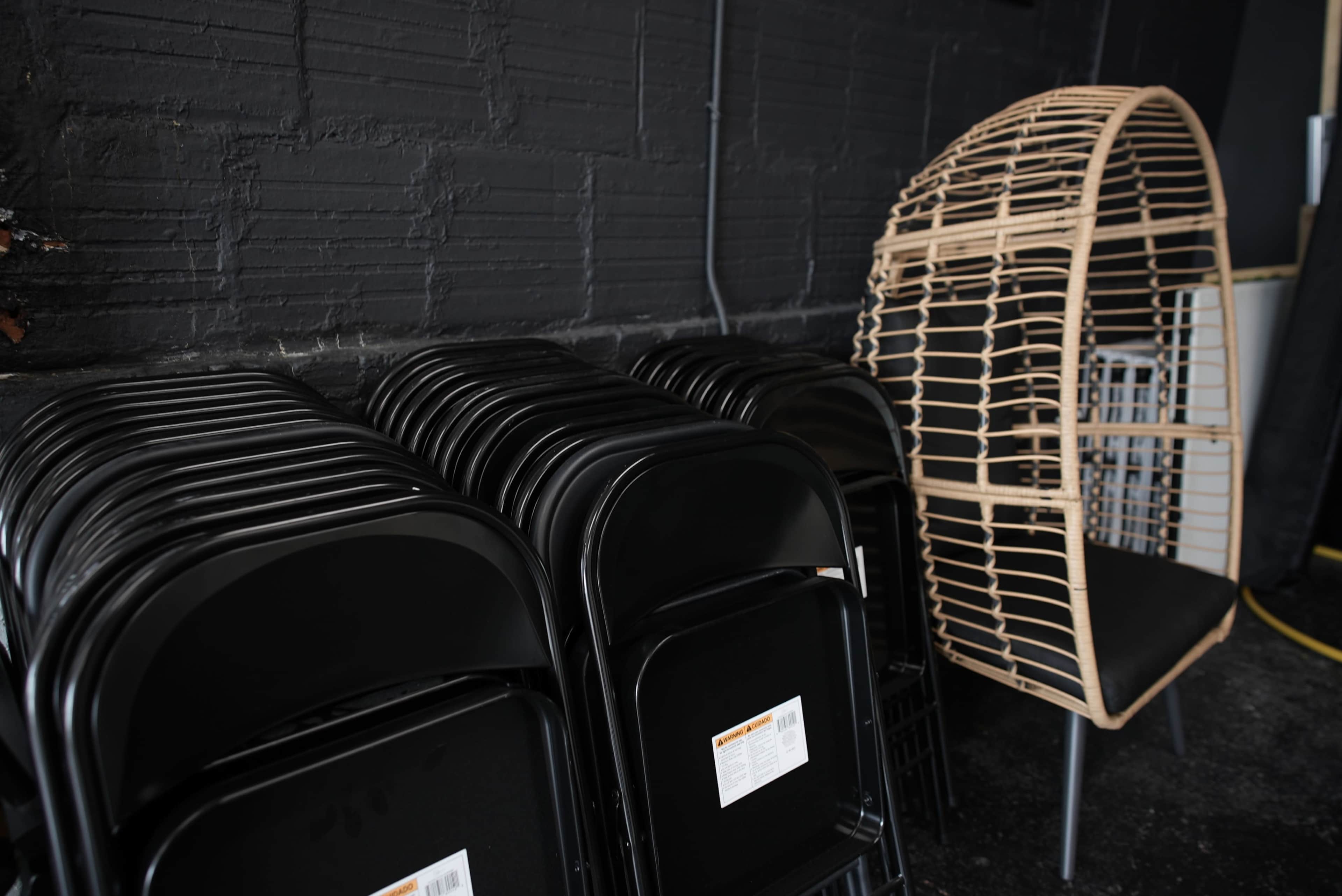 A stack of black folding chairs beside a large wicker chair against a dark wall.