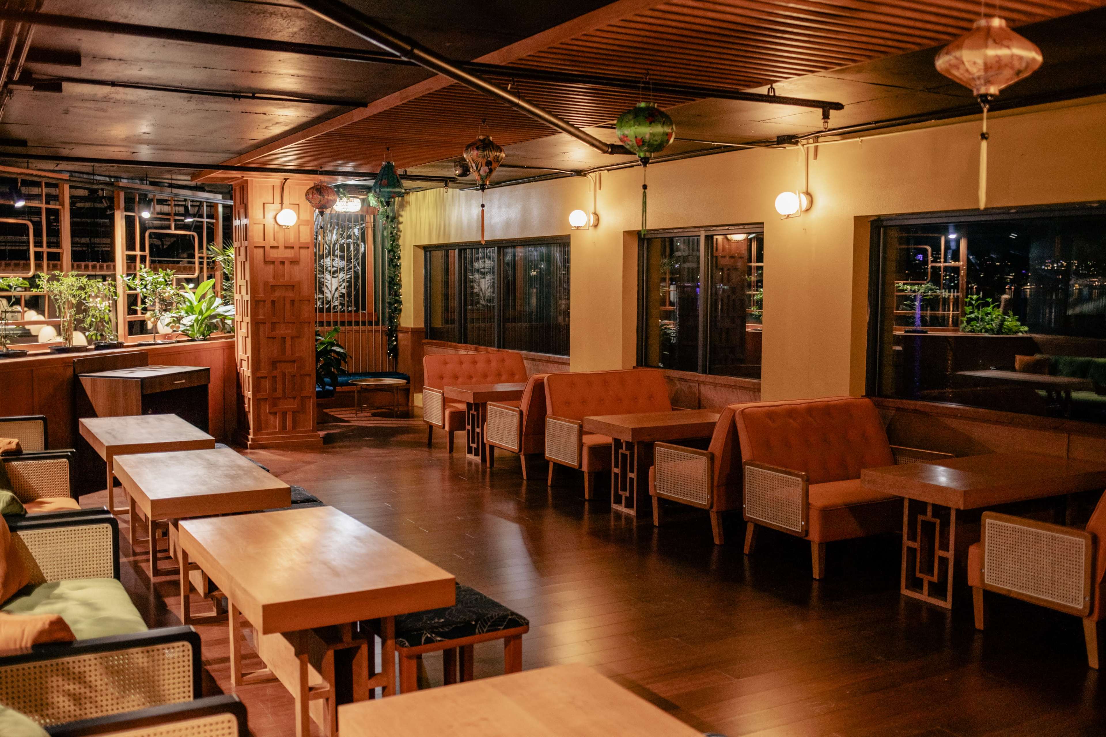 The image shows a spacious restaurant interior with wooden tables and seating, illuminated by pendant lights and decorated with plants.
