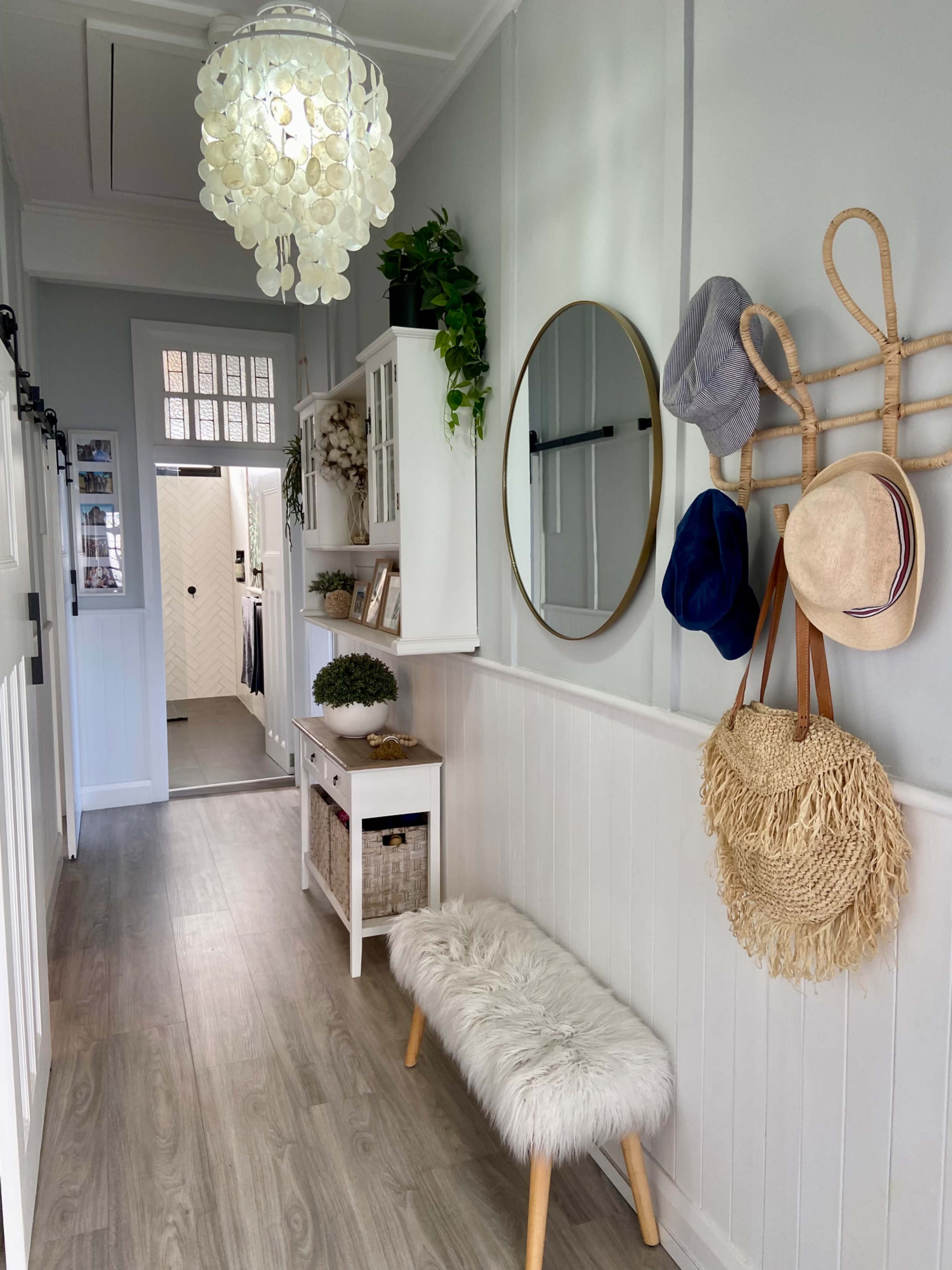 The image shows a narrow hallway featuring a white bench with a fluffy cushion, a round mirror on the wall, various hats hanging on hooks, and a light fixture above.
