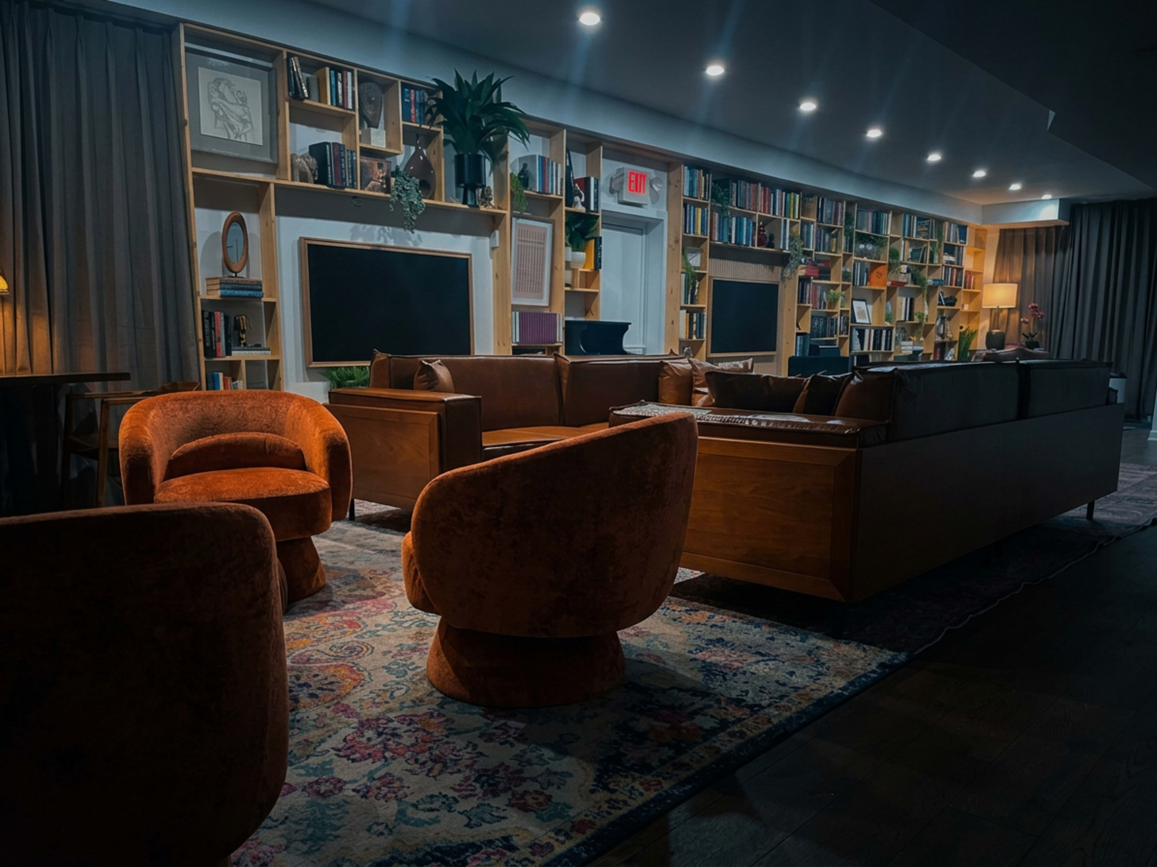 The image shows a cozy lounge area with two orange velvet chairs, a wooden sofa, and a bookshelf filled with books against a wall.