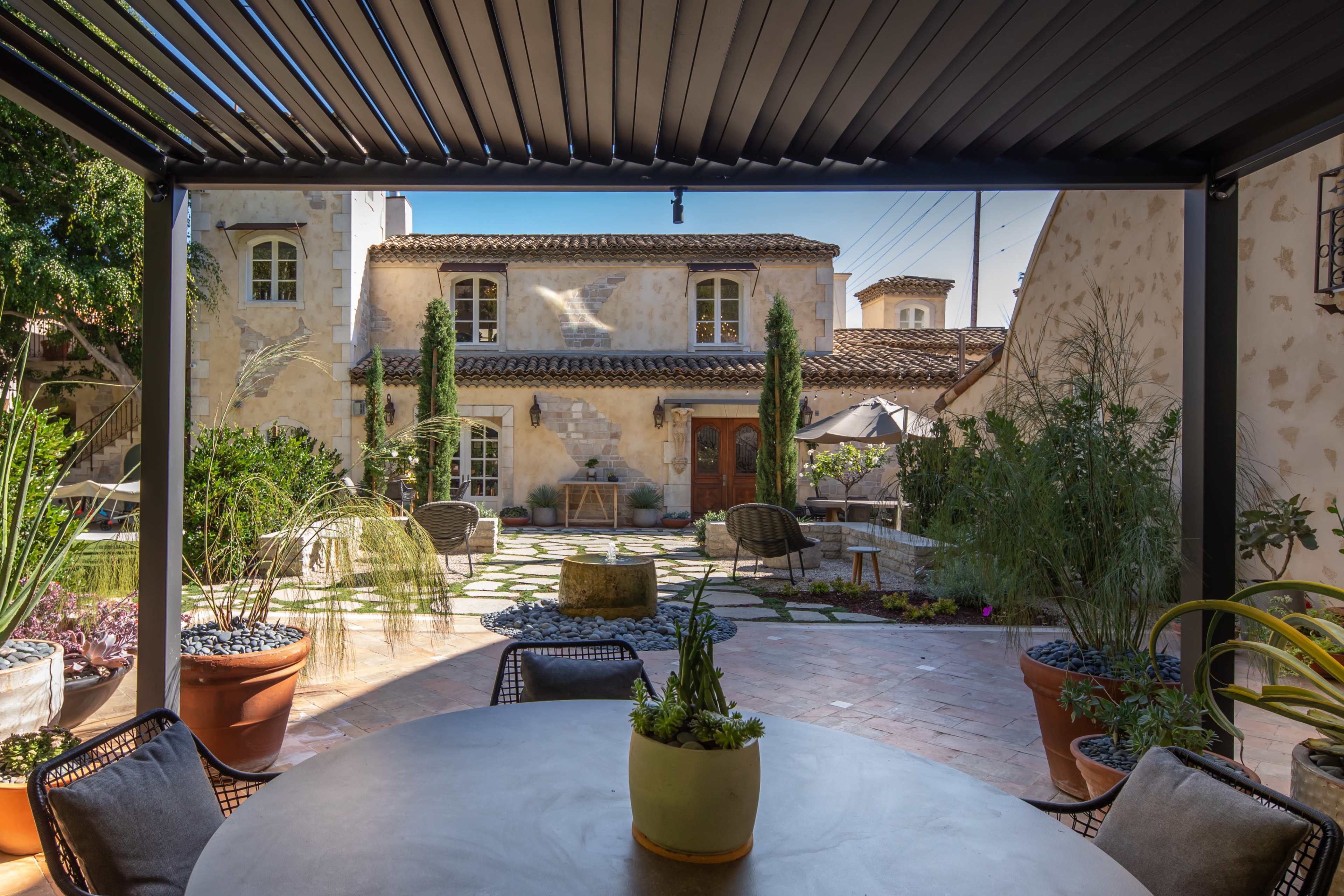 A patio with a round table and potted plants overlooks a courtyard featuring a stone pathway, greenery, and a rustic house in the background.