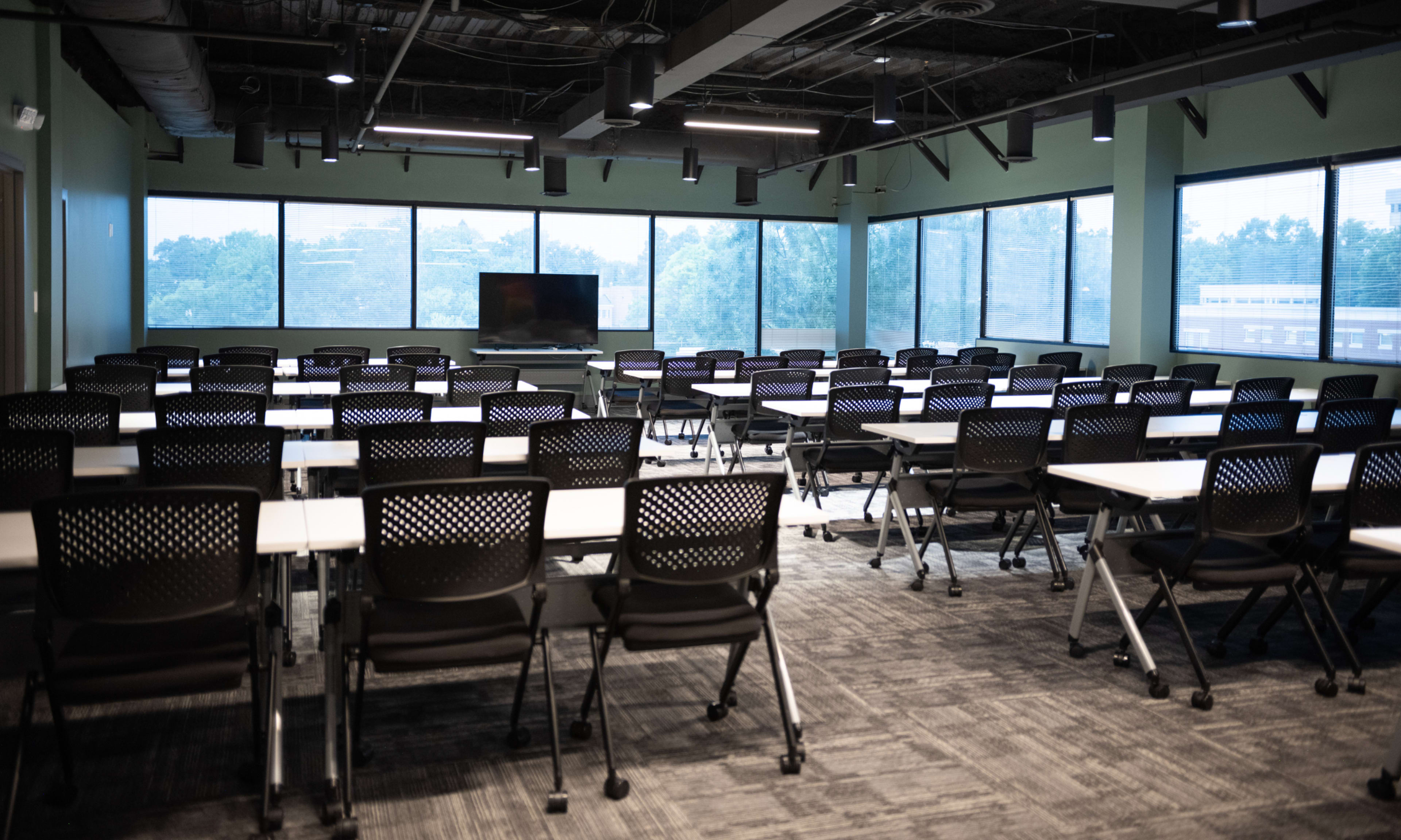 A neatly arranged meeting room features rows of black chairs and tables, with large windows allowing natural light to illuminate the space.