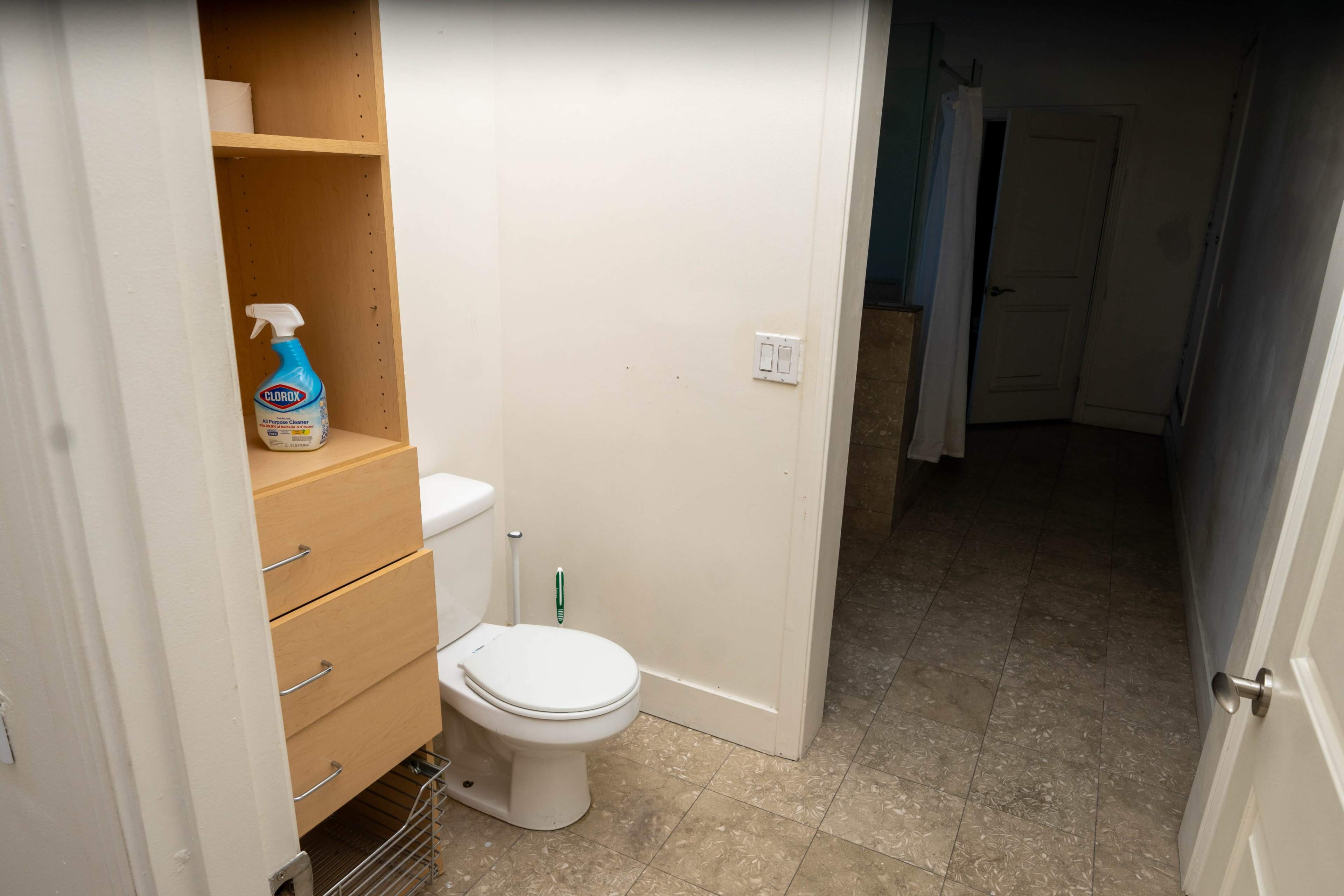 A bathroom with a toilet, a small shelving unit, and a doorway leading to another room.