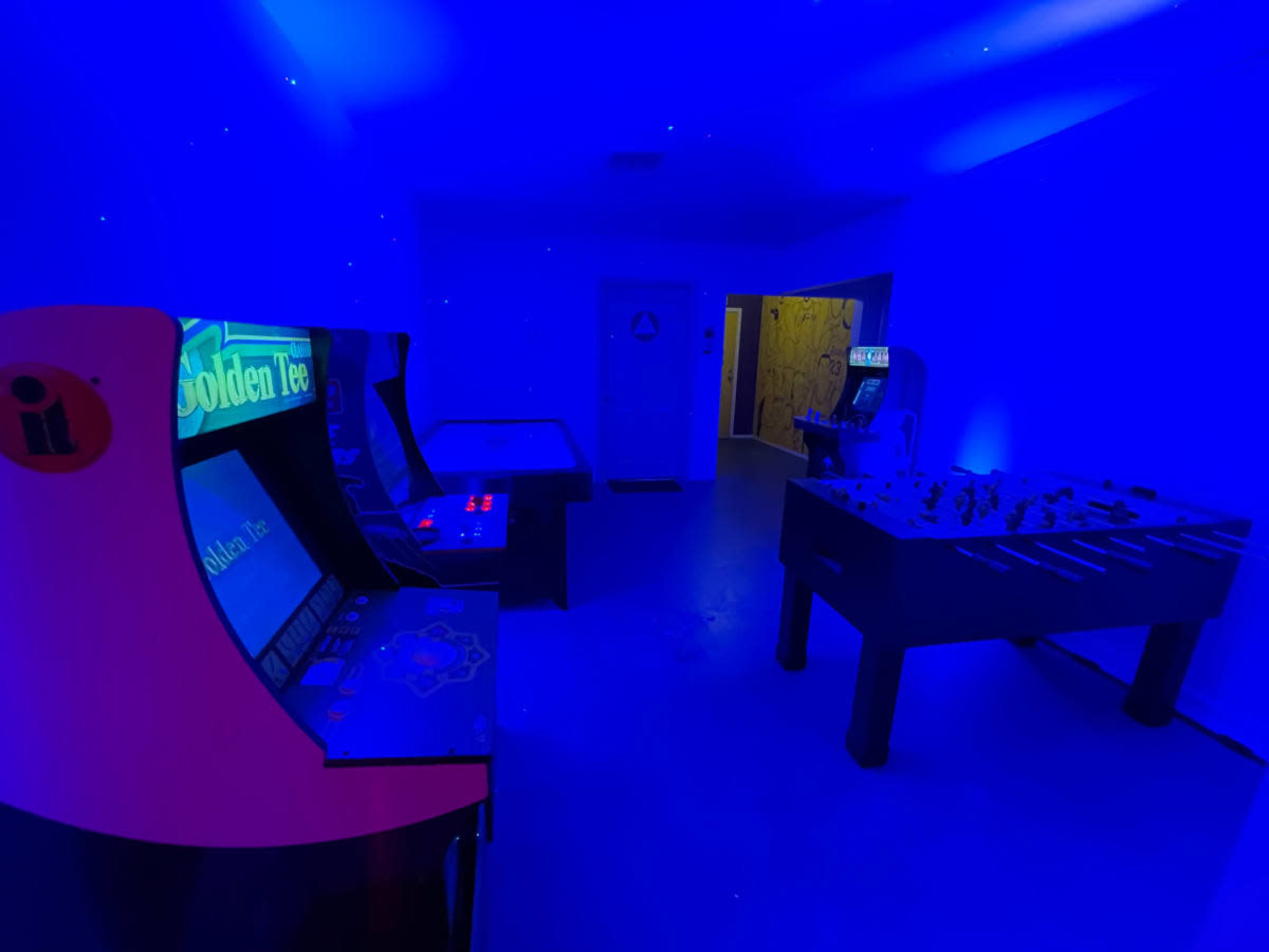 The image shows a dimly lit arcade room with blue lighting, featuring classic arcade machines and a foosball table.