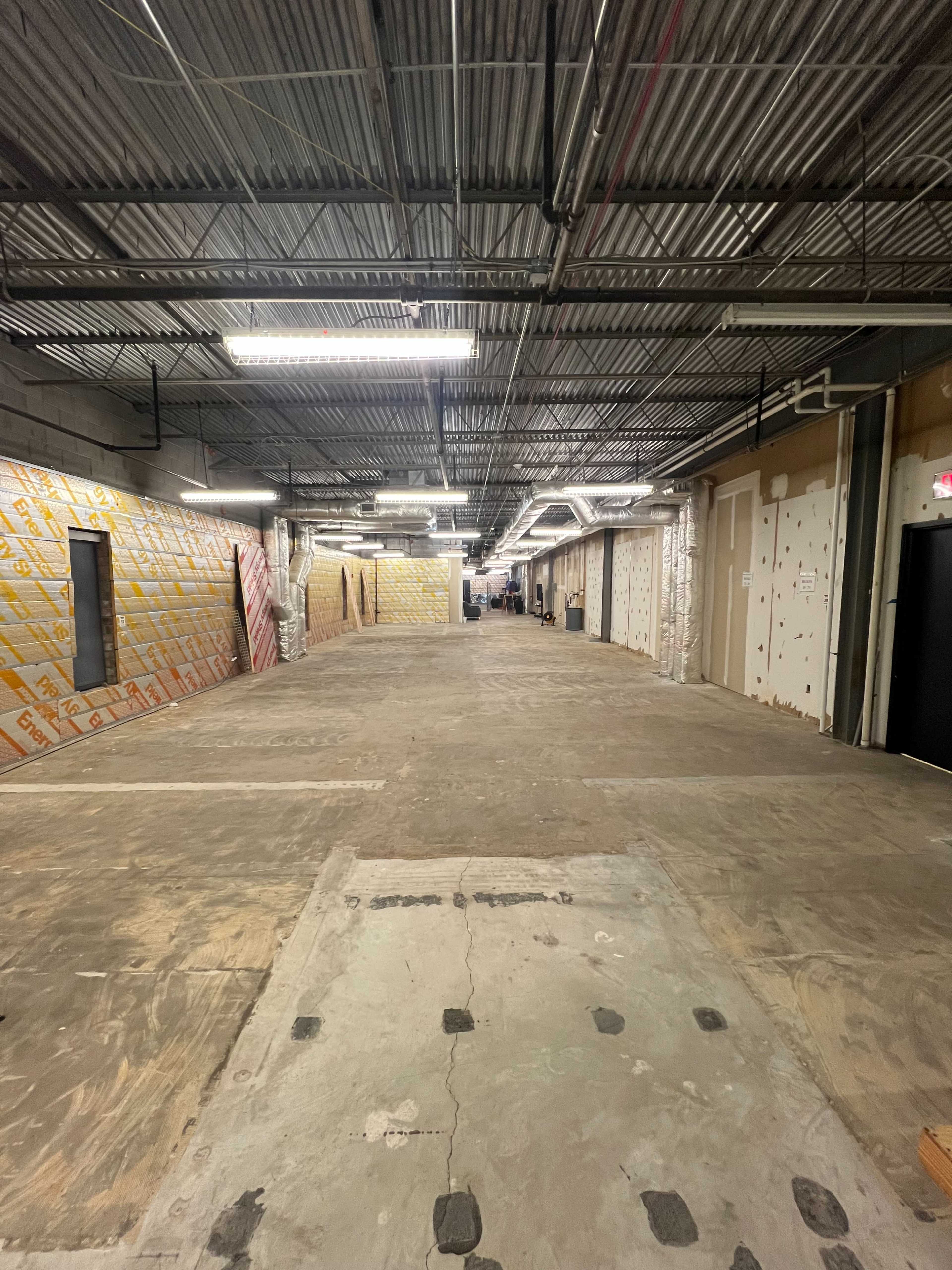 The image shows a long, empty corridor with exposed walls and unfinished flooring, illuminated by fluorescent lights.