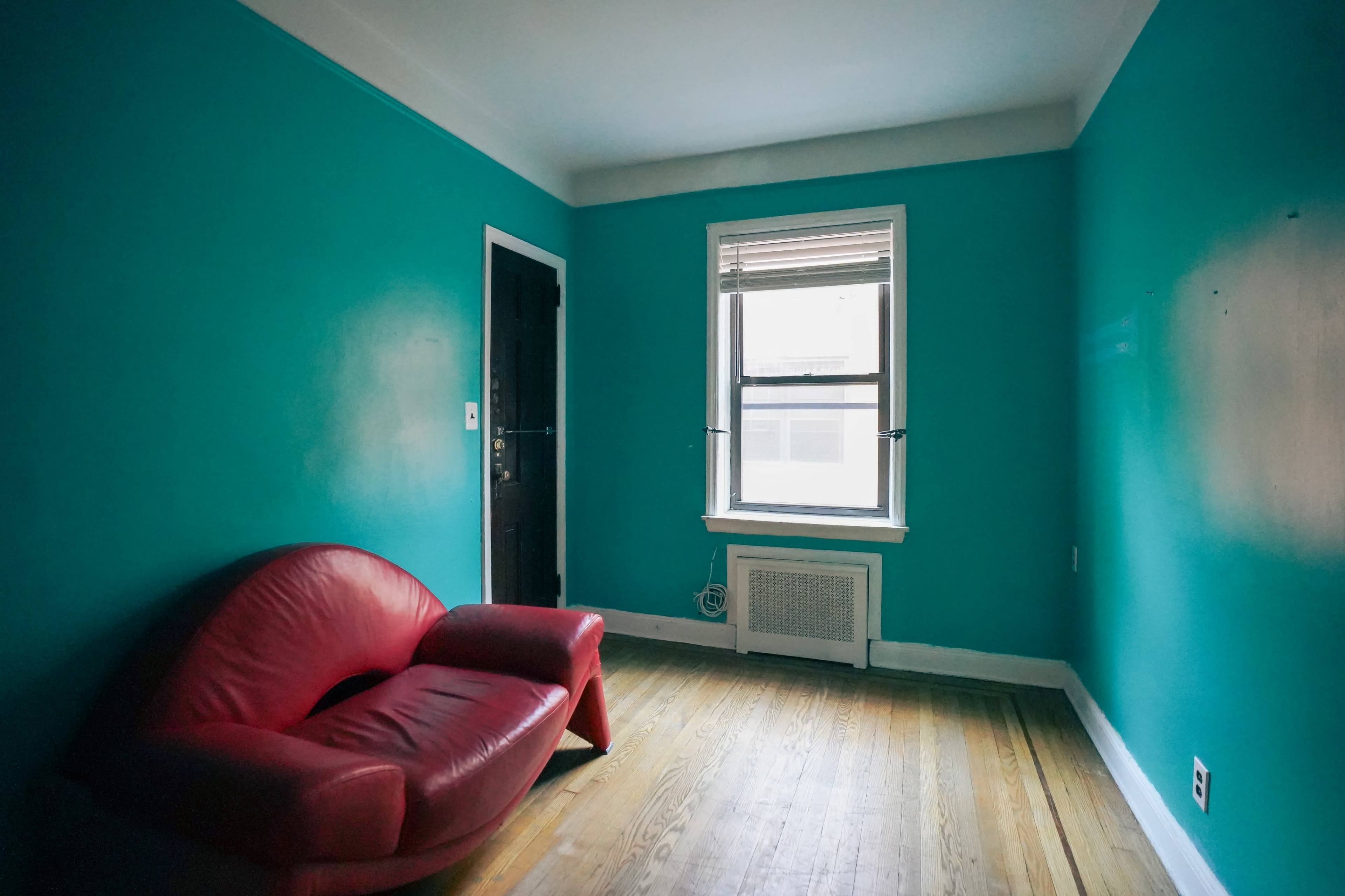 The image shows a small room painted turquoise with a red armchair, a window, and hardwood flooring.