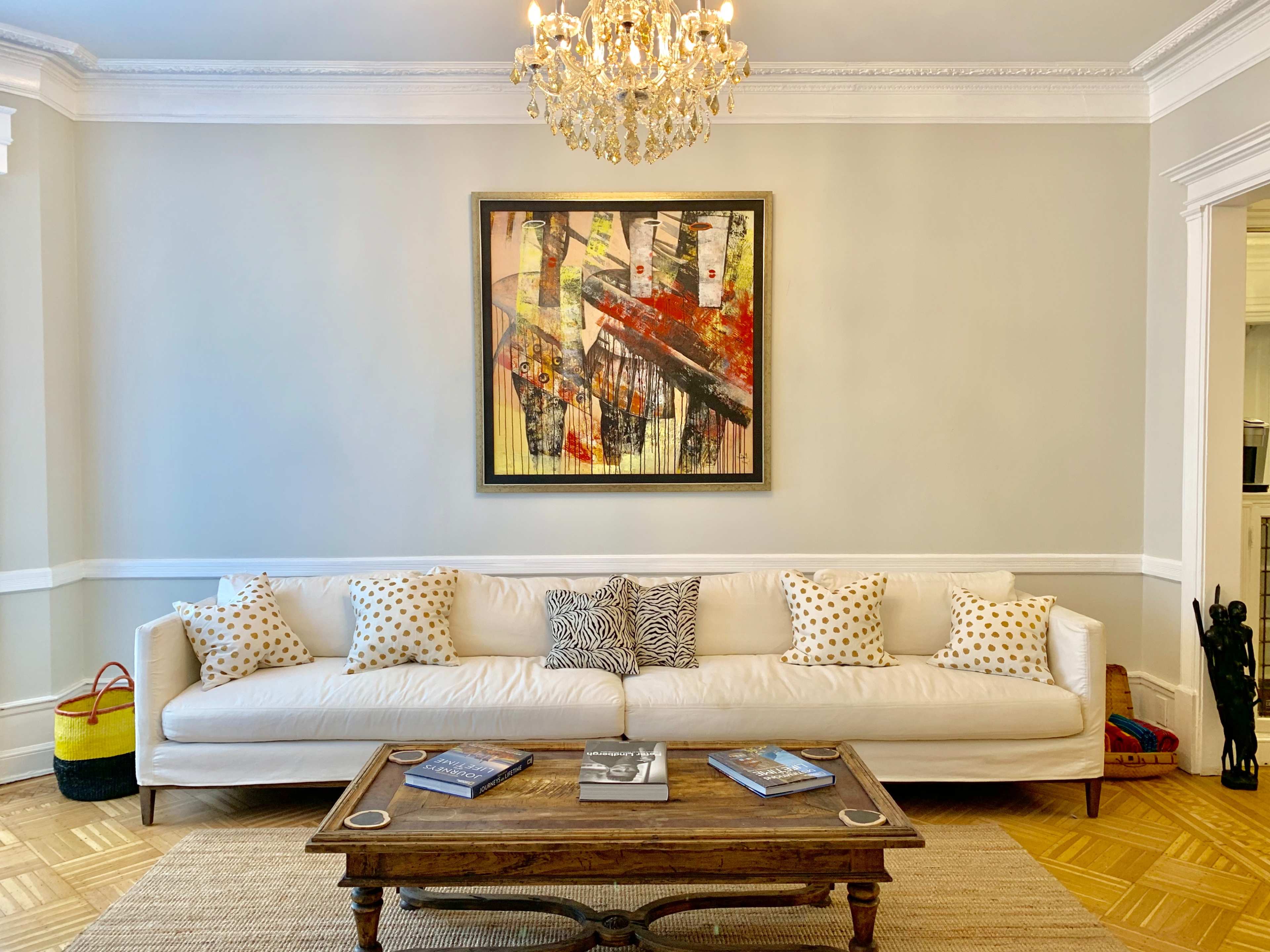 A white leather couch with decorative pillows is positioned in front of a wooden coffee table, under a crystal chandelier, against a light gray wall featuring an abstract painting.