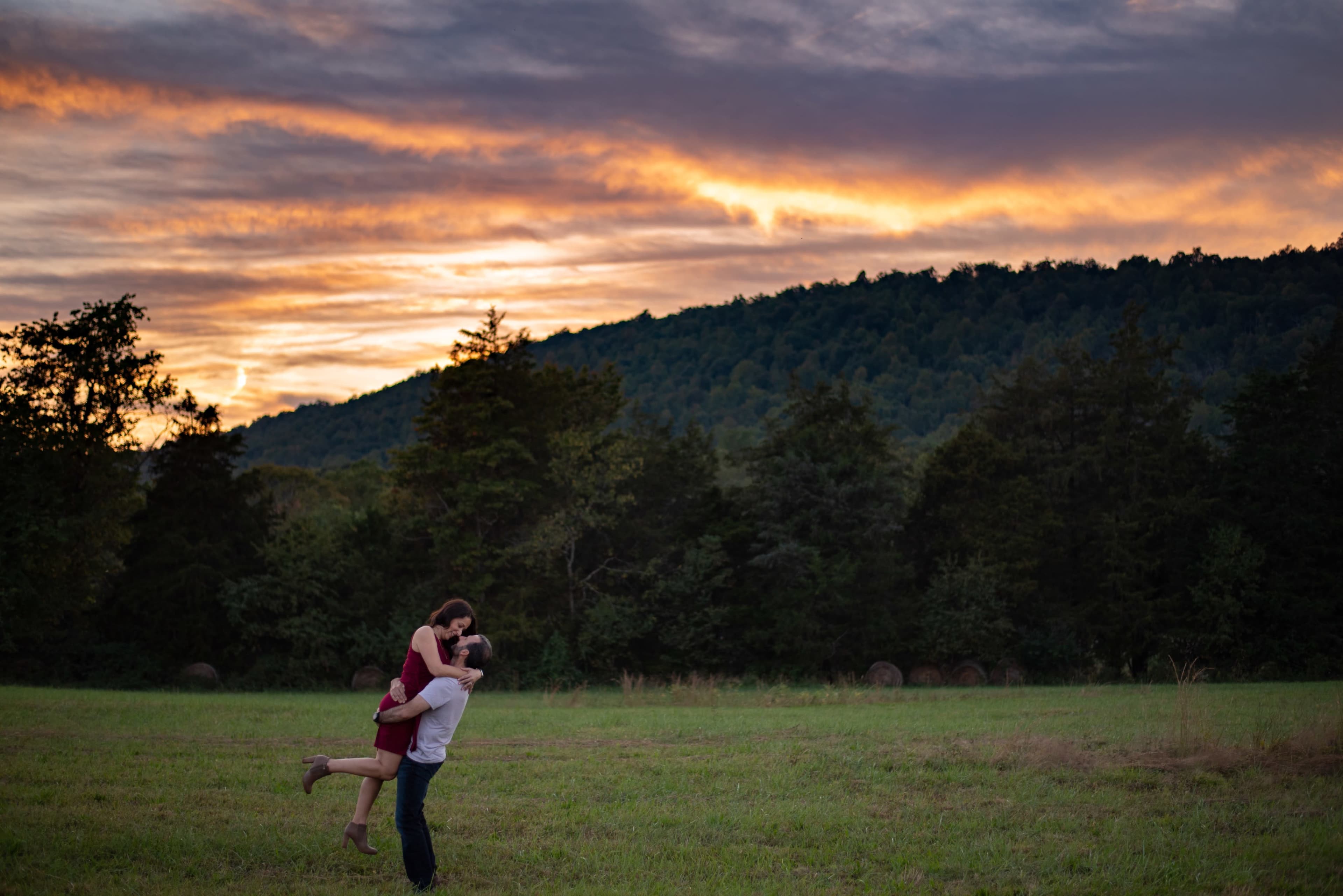 A couple embraces in a field at sunset, surrounded by trees and rolling hills.