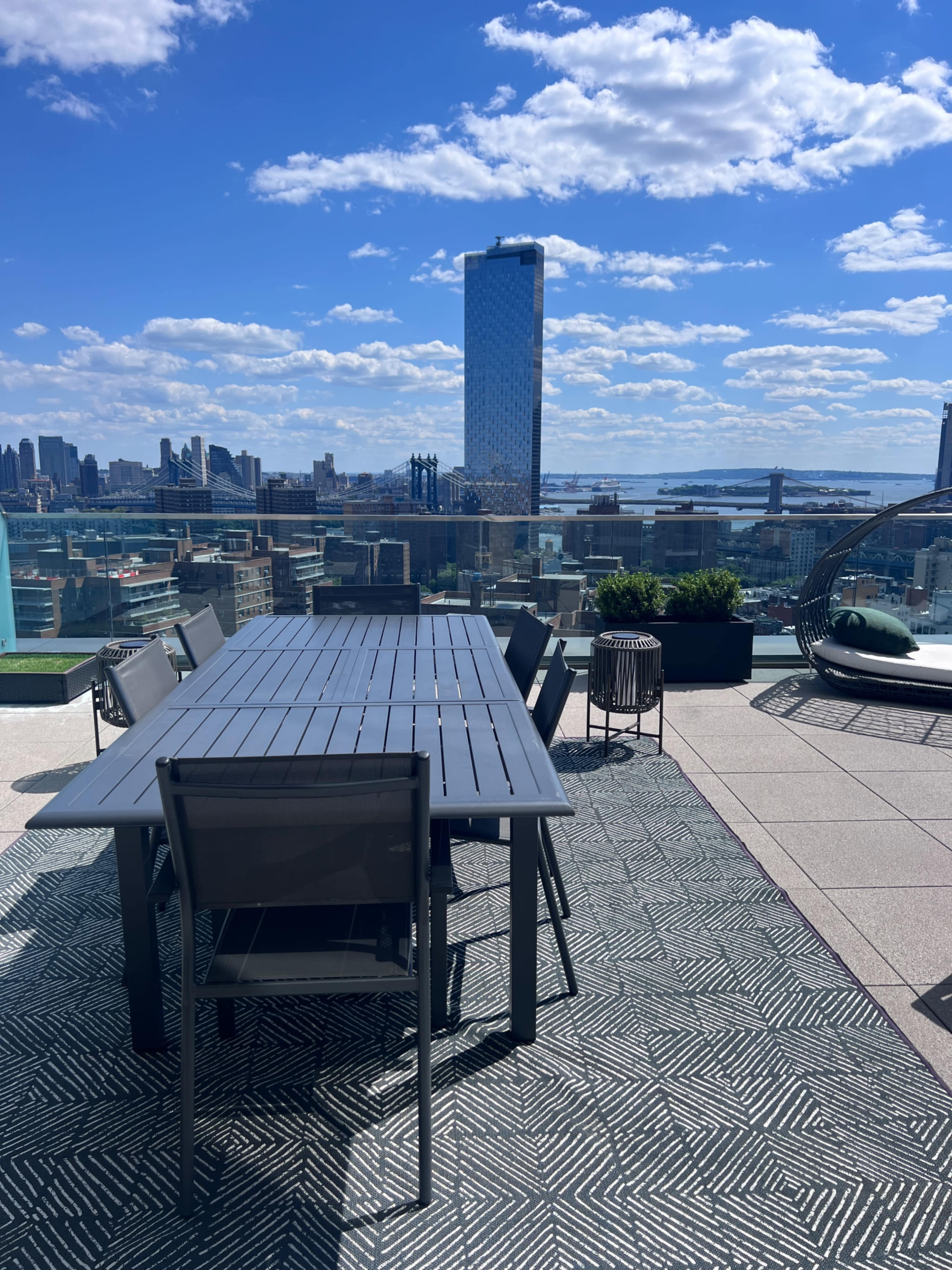 A rooftop terrace features a large dining table with chairs, overlooking a city skyline and a body of water under a partly cloudy sky.