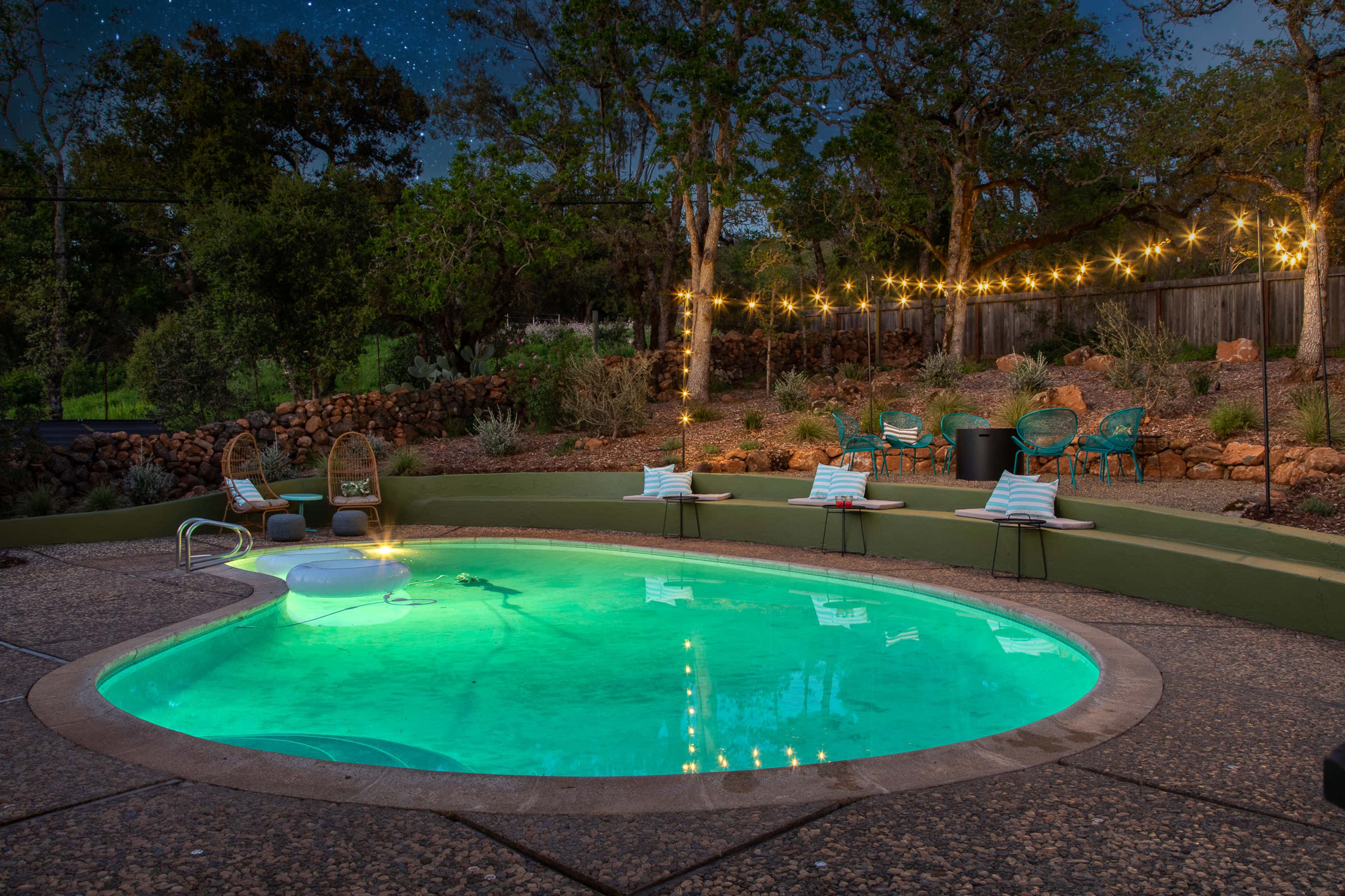 A illuminated swimming pool is surrounded by seating areas and decorative lights in a landscaped backyard at dusk.