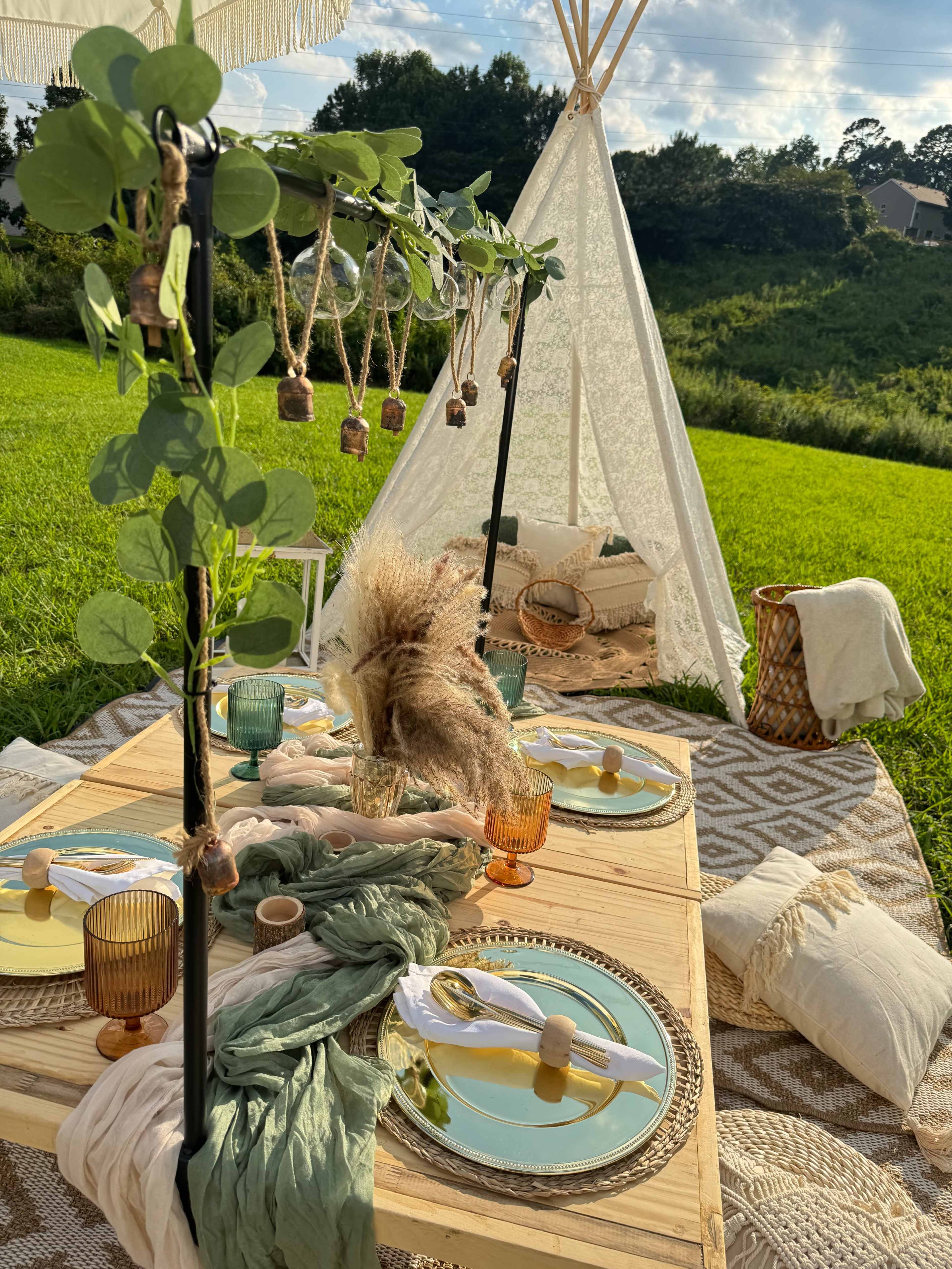 The image shows a decorated outdoor dining setup with a wooden table, colorful glassware, and a teepee in a grassy area.