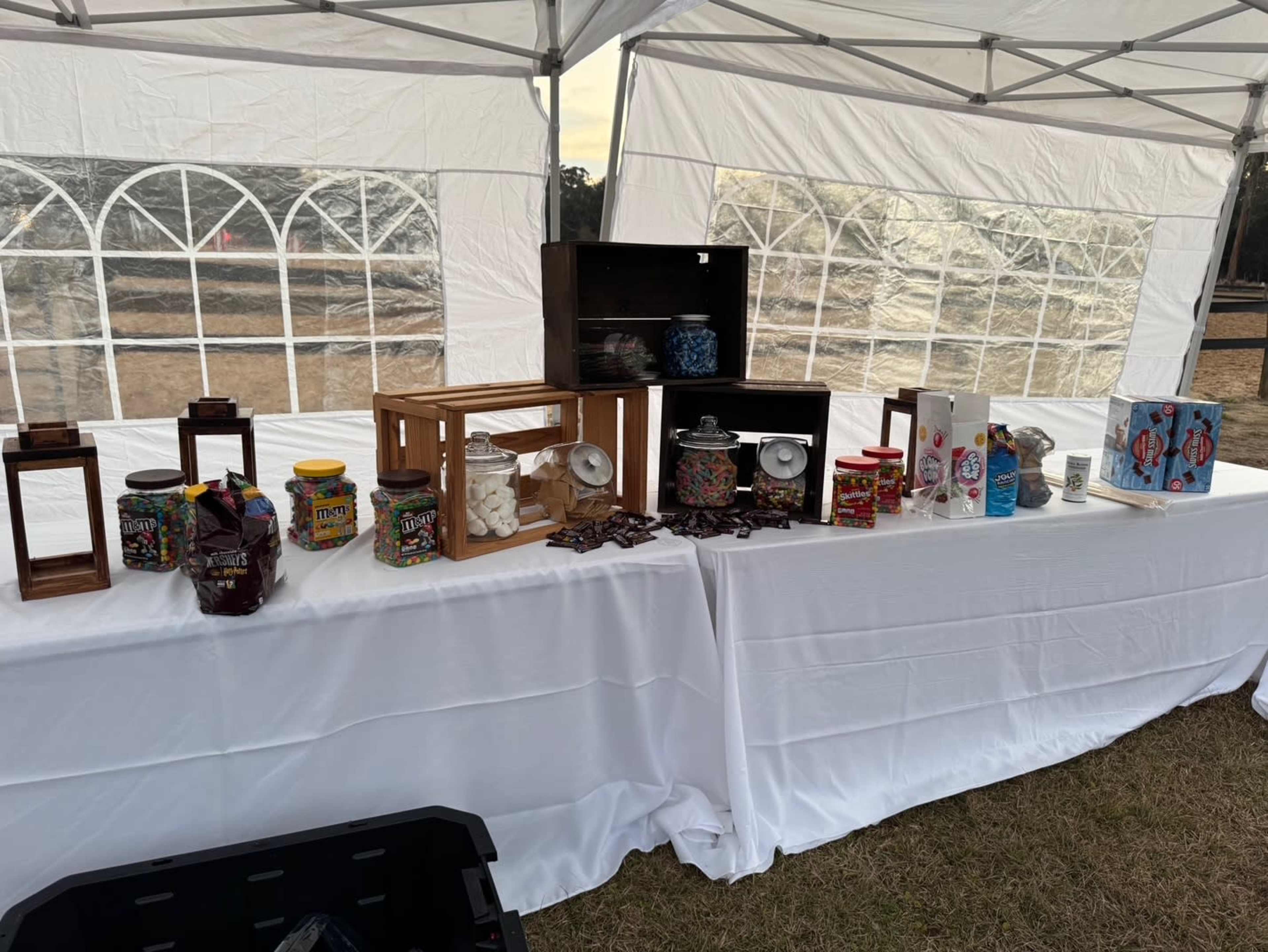 A table covered with a white cloth displays various jars and containers filled with colorful candies and snacks under a tent.