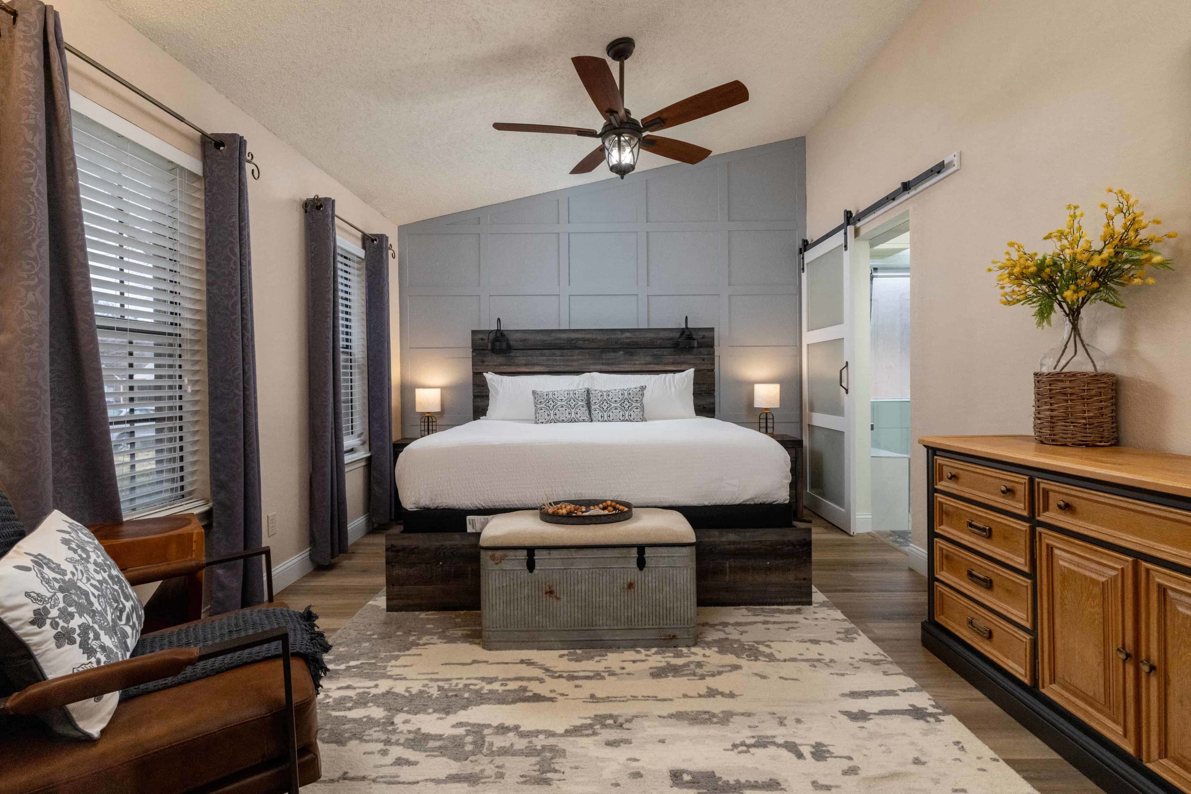 The image shows a cozy bedroom featuring a large bed with a decorative headboard, a wooden dresser, and a rug on the floor, with natural light coming through the curtains.