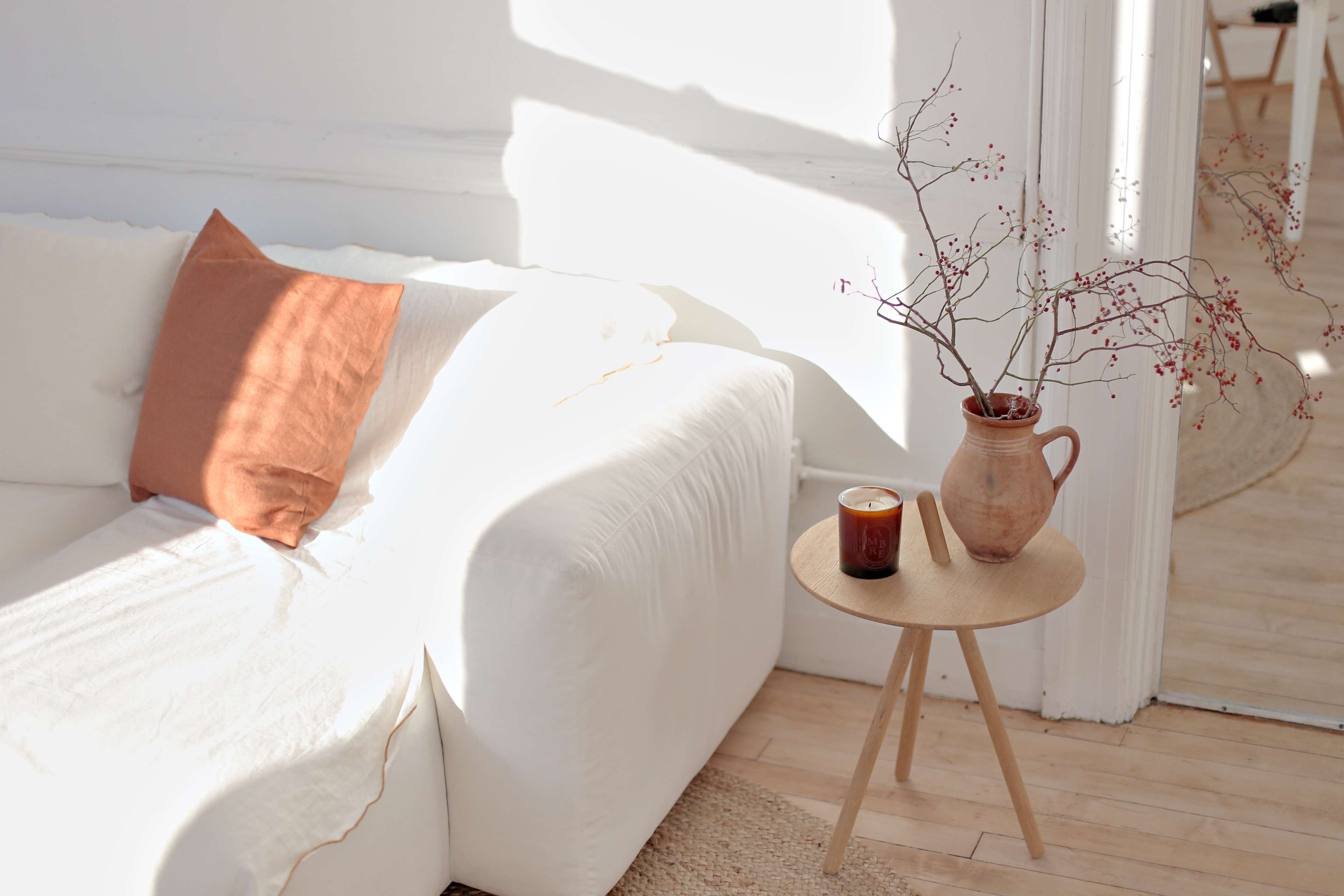 A white sofa with a rust-colored pillow sits beside a wooden side table holding a dark drink and a vase with branches.