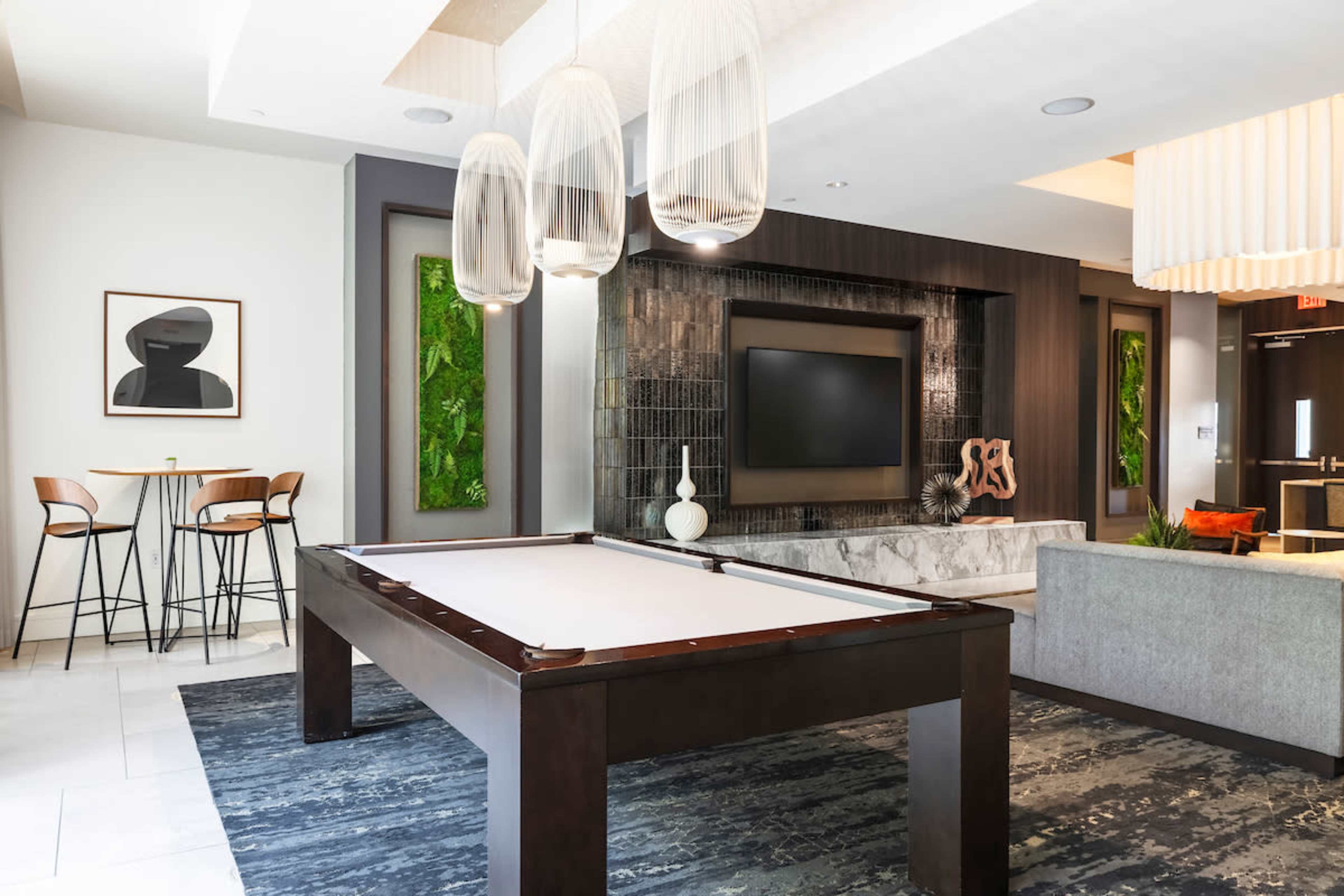 The image shows a modern lounge area featuring a billiard table, minimalist furniture, and decorative wall features, along with a large television and stylish lighting fixtures.