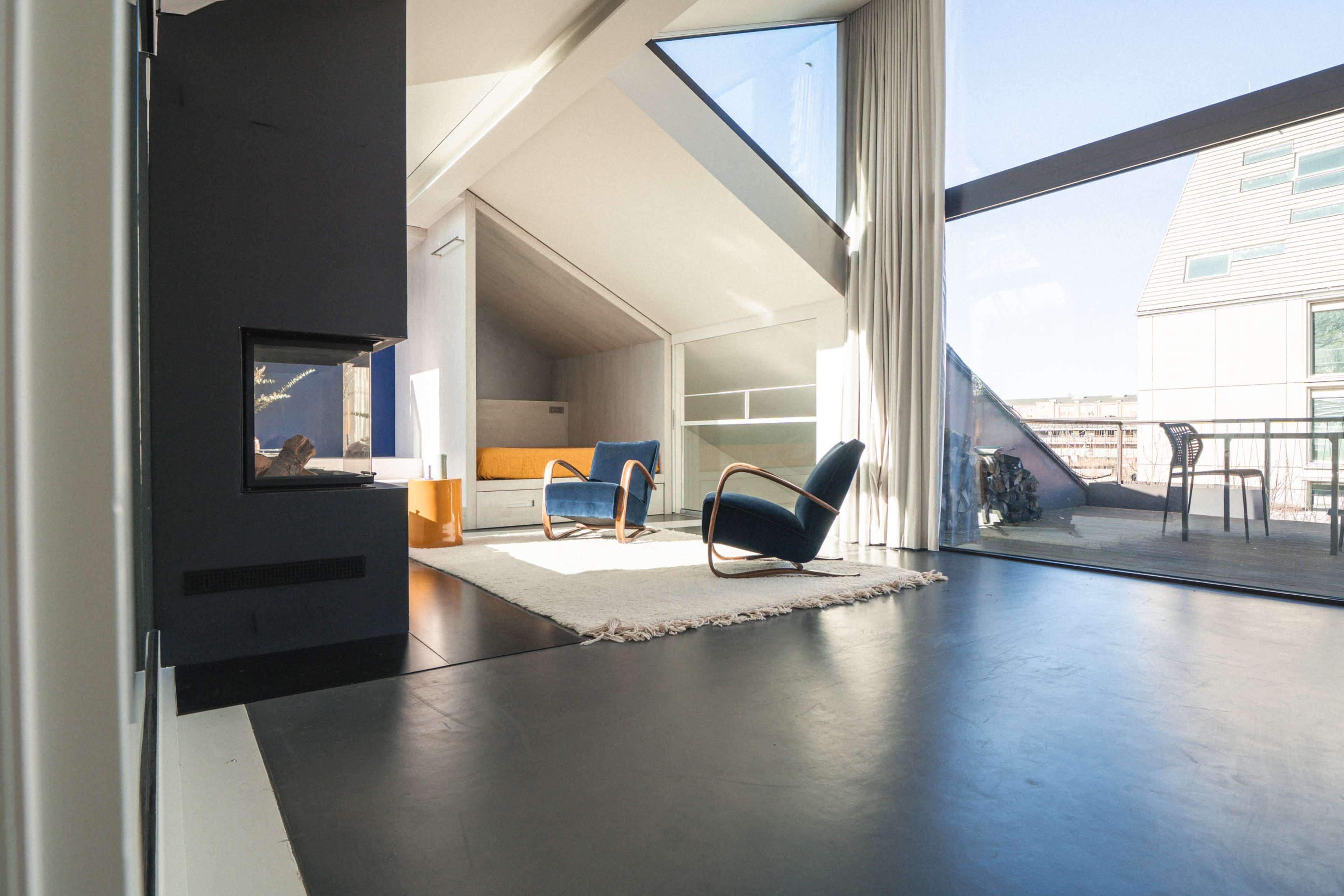 The image shows a modern living room with a fireplace, two blue chairs, a large window, and an expansive view of the outdoors.