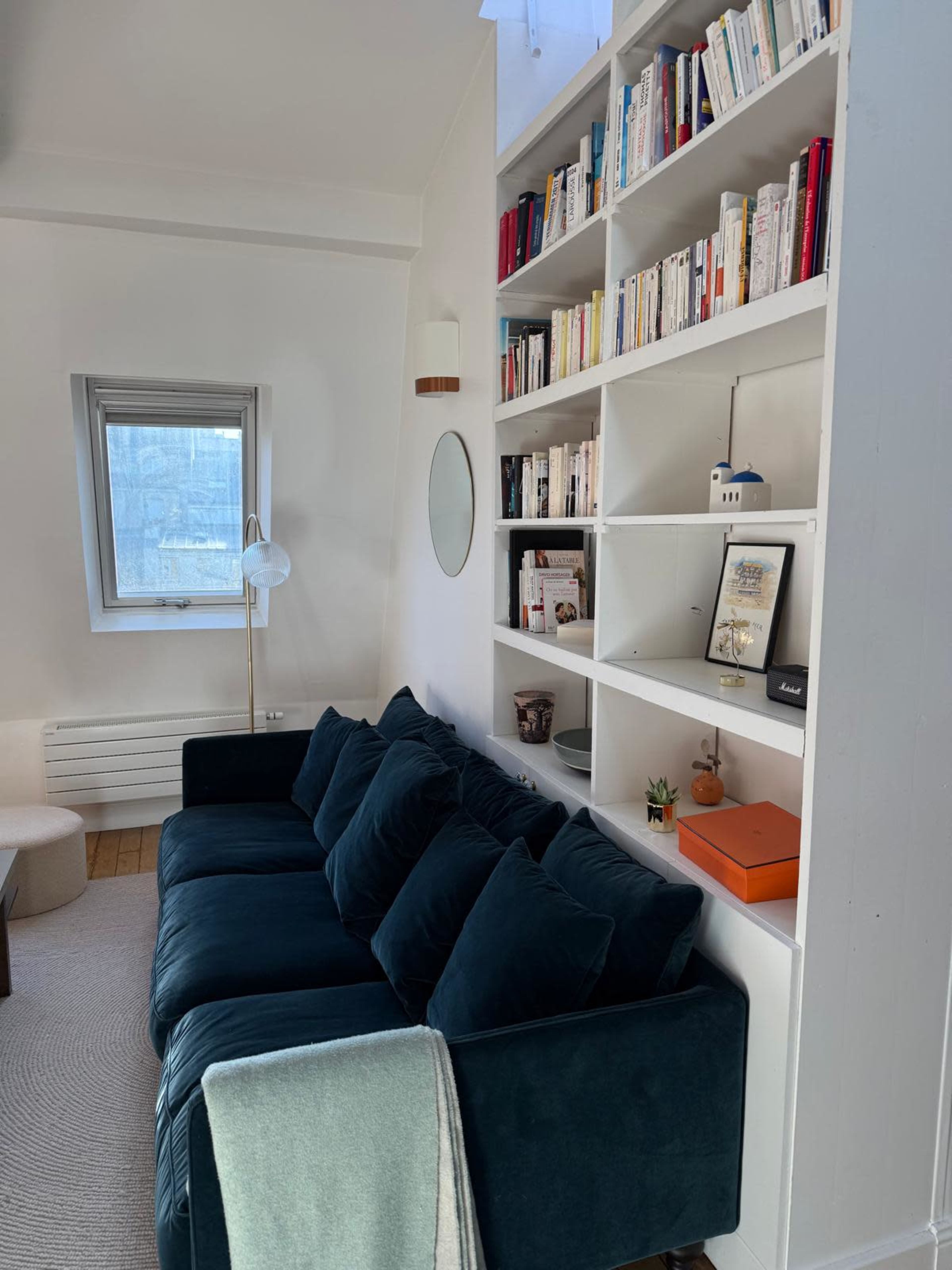 The image shows a cozy living room featuring a dark blue sofa, a bookshelf filled with books, and a circular mirror on the wall.