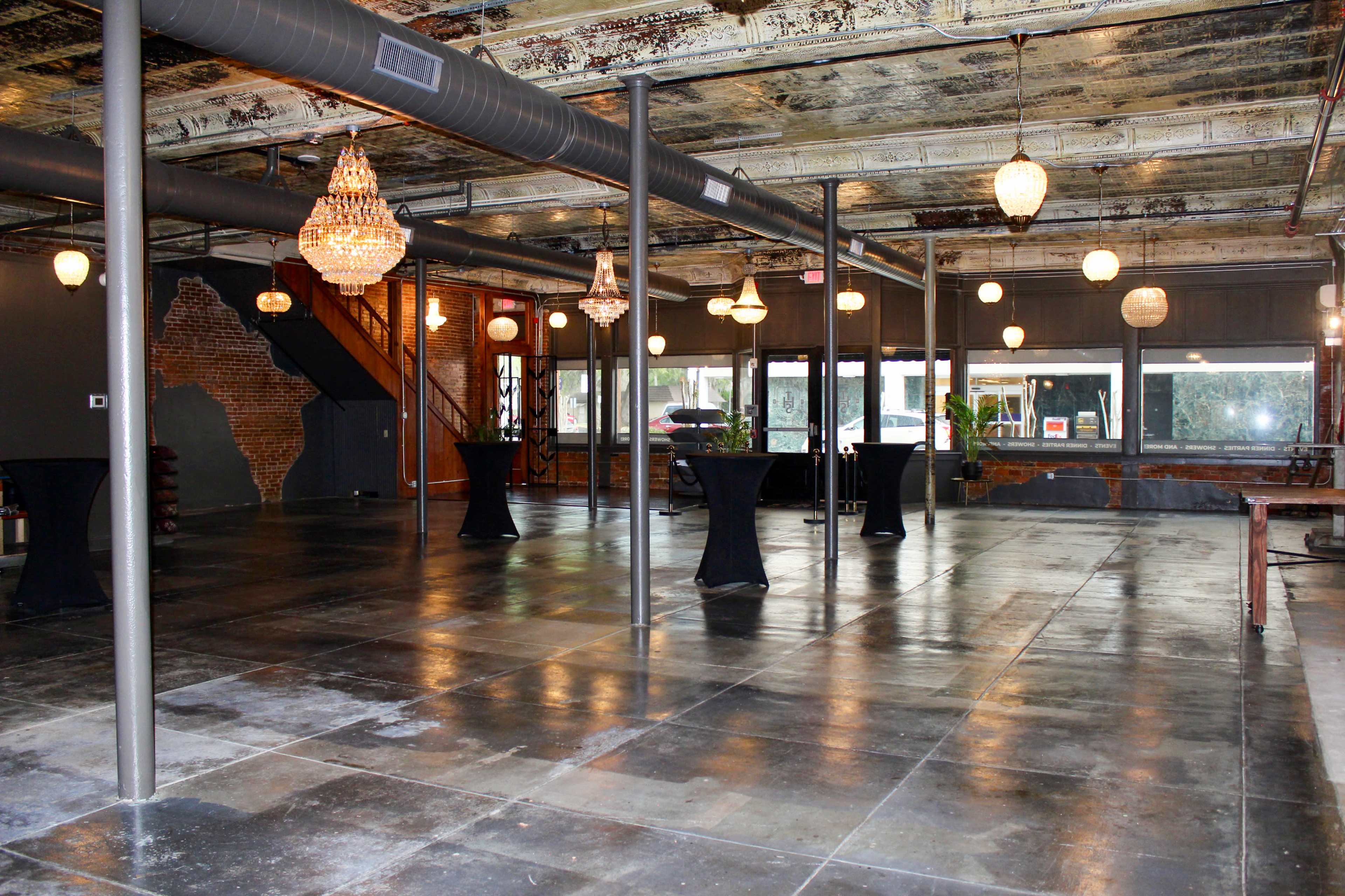 The image shows a spacious industrial-style room with exposed brick walls, a polished concrete floor, hanging light fixtures, and several tall tables set in a clean, open layout.