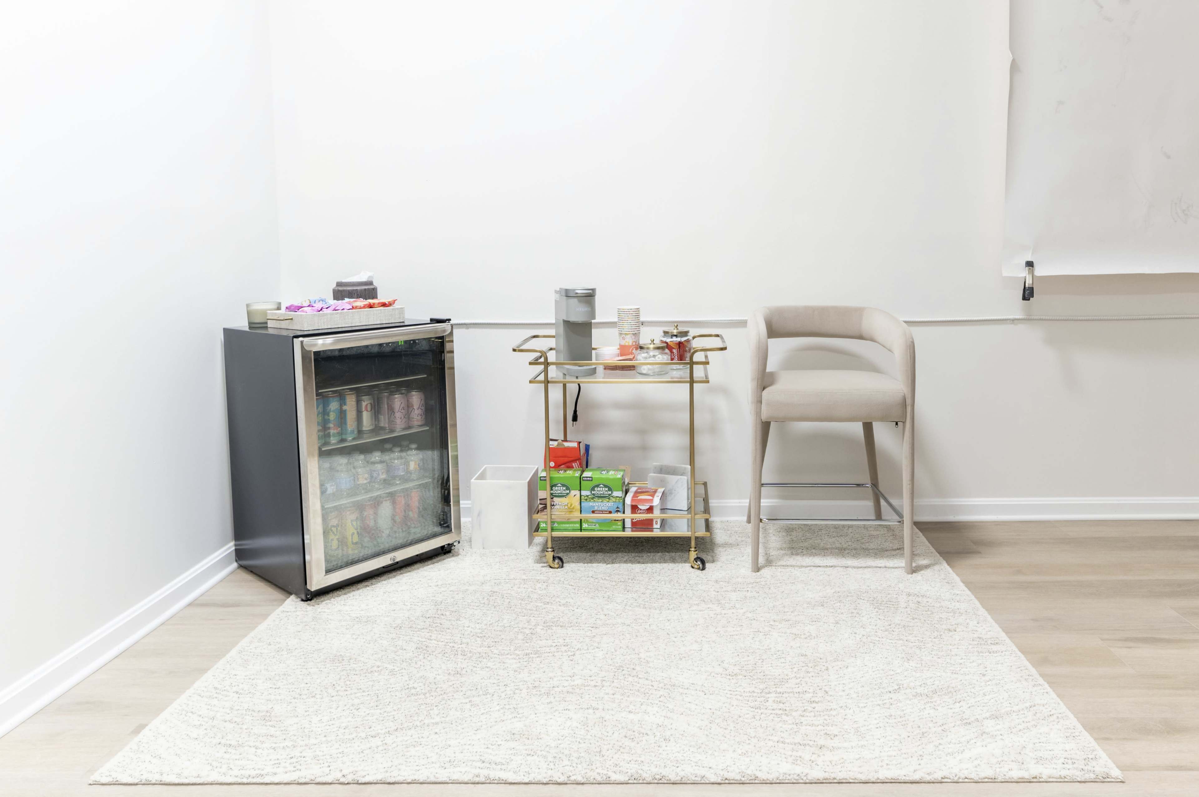 A small, minimalistic corner features a black mini fridge filled with drinks, a gold bar cart displaying snacks, and a light-colored chair on a textured rug.