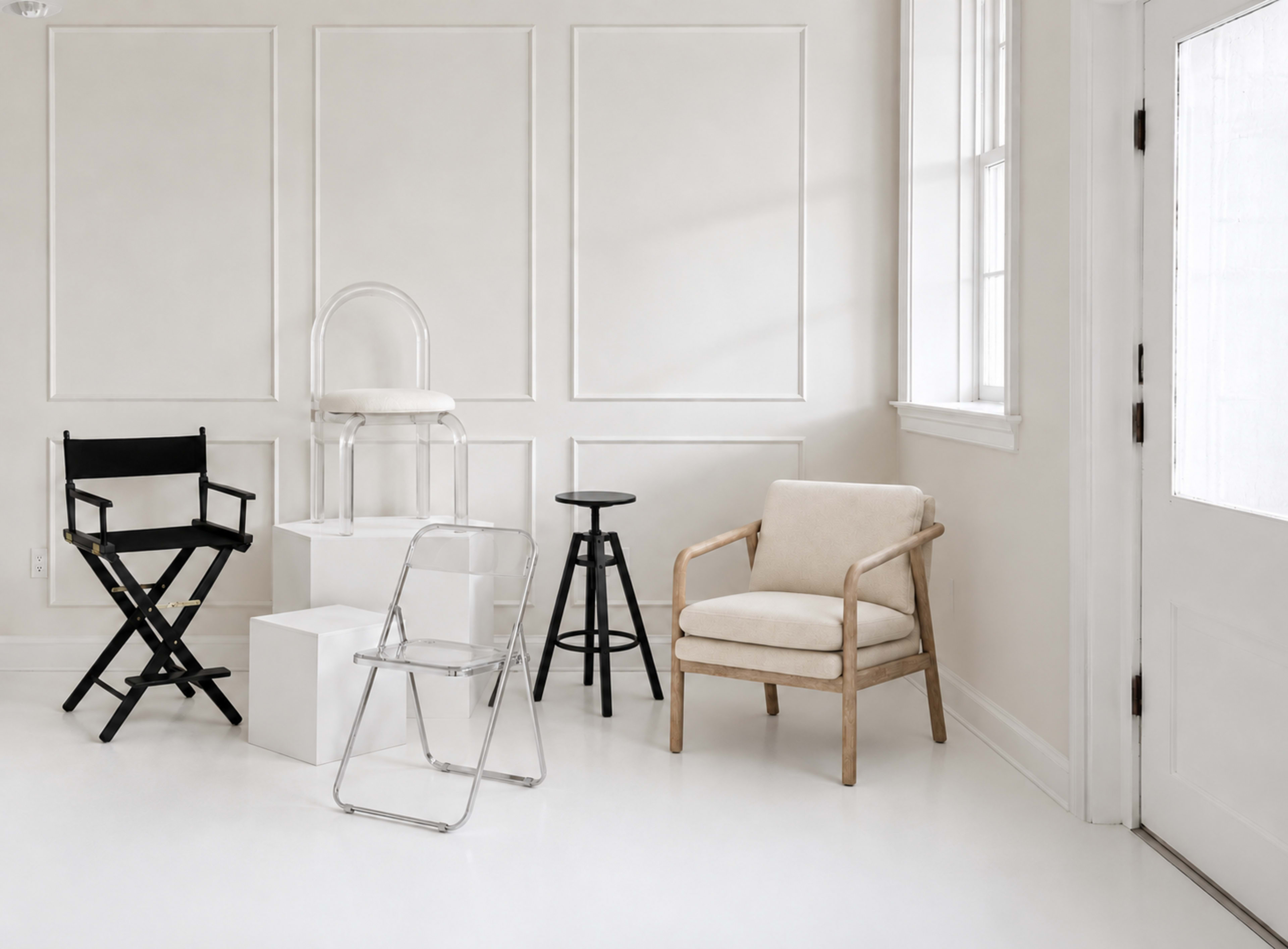 The image shows a modern, minimalist interior featuring a variety of chairs, including a black director's chair, a transparent chair, a wooden armchair, a metal stool, and a folding chair, arranged in a light, neutral-colored room with wainscoting and a window.