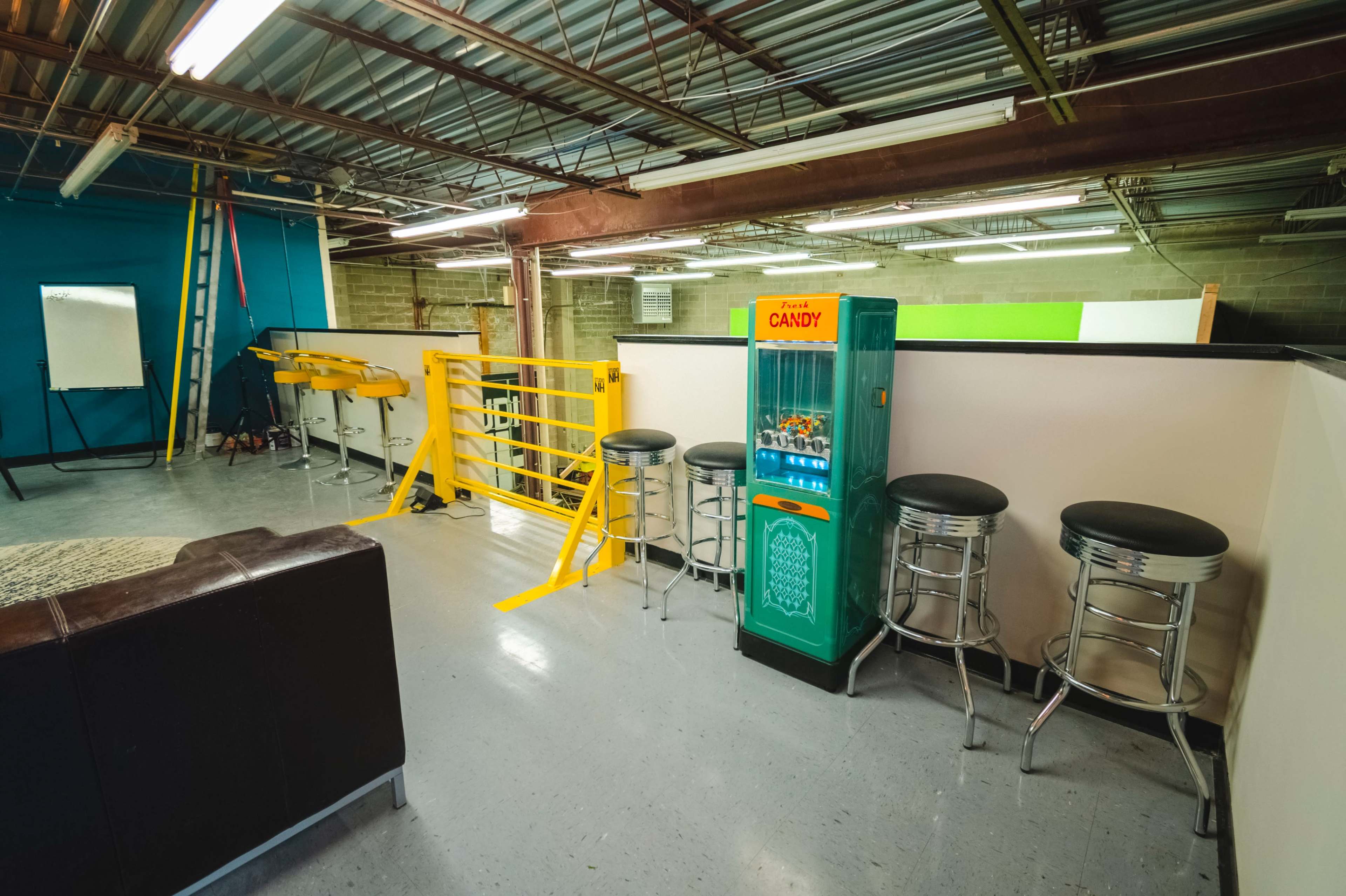 An interior space featuring a candy vending machine beside several metal stools and a yellow safety railing.