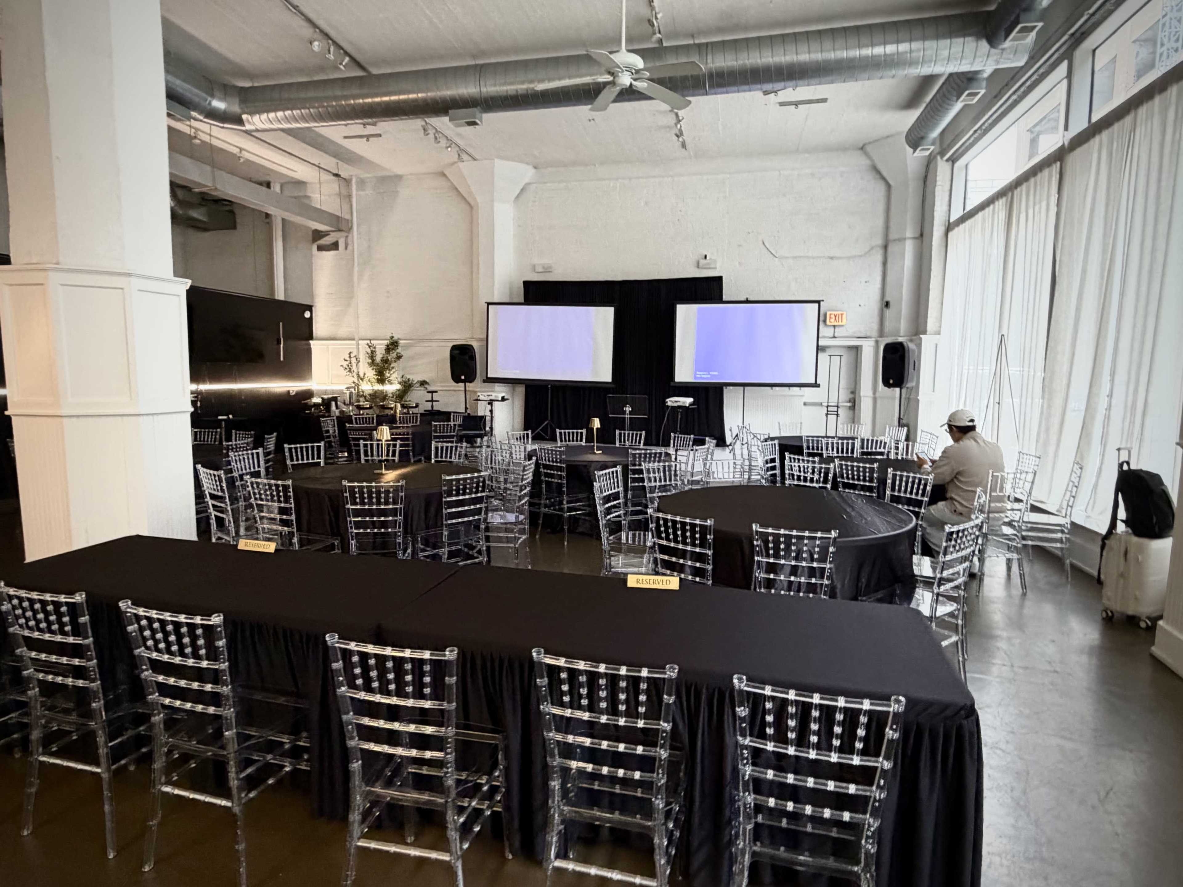 The image shows a spacious event venue arranged with several round tables and a long rectangular table, featuring two screens and a dark backdrop.