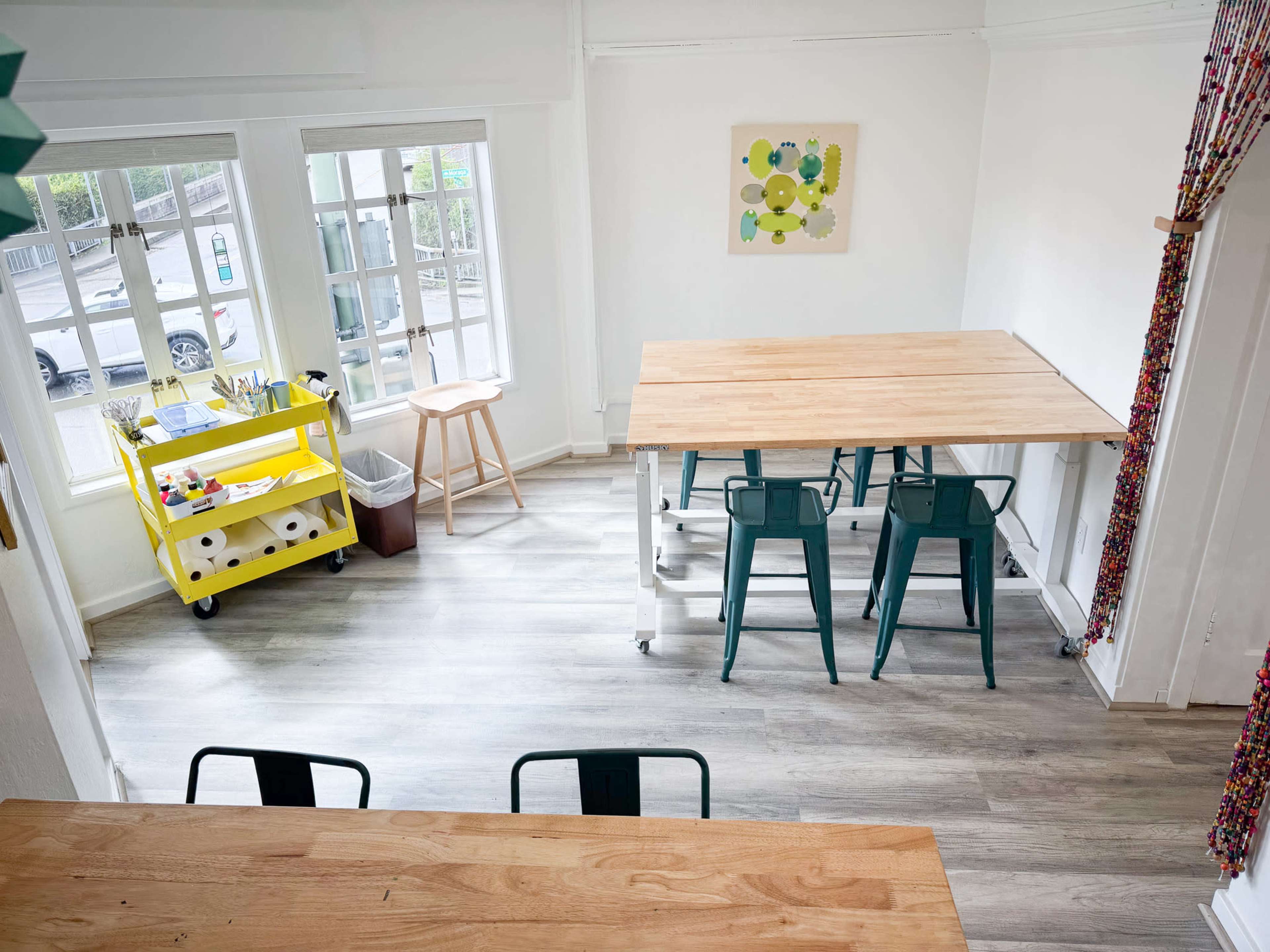 A bright, modern workspace featuring two tables, green chairs, a rolling cart with supplies, and large windows allowing natural light.