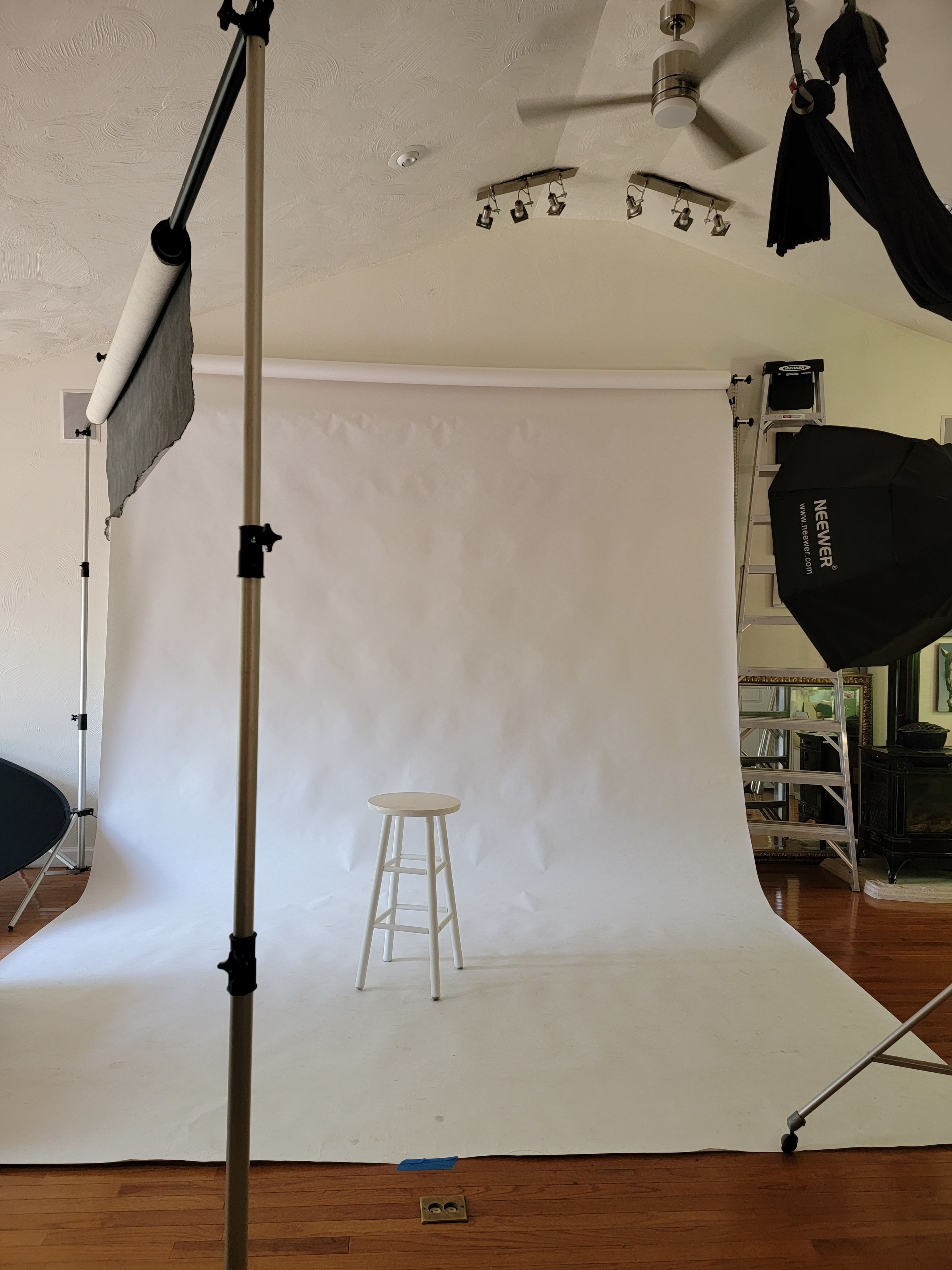 The image shows an empty photography studio with a white backdrop and a single stool positioned in front of it.
